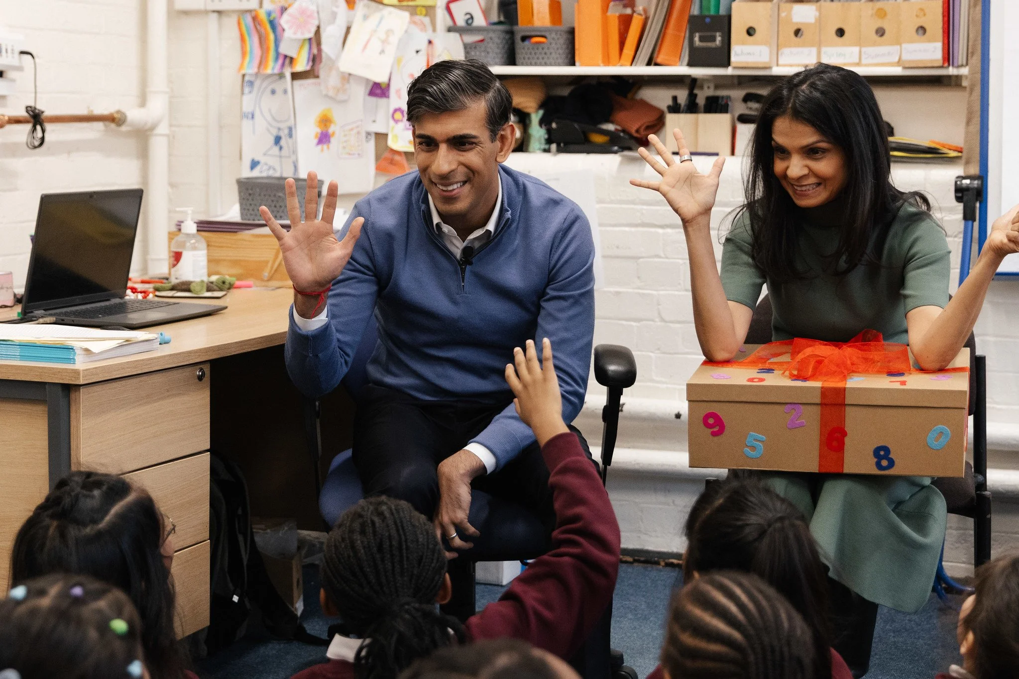 Adults engaging with children in a classroom, sitting at a table surrounded by young students. The man has dark hair, wears a blue sweater, and smiles while waving. The woman has dark hair, wears a green top, and also waves. Children are reaching up to high-five with the adults. The classroom has shelves with binders, artwork on the wall, and a decorated box with numbers and a bow.
