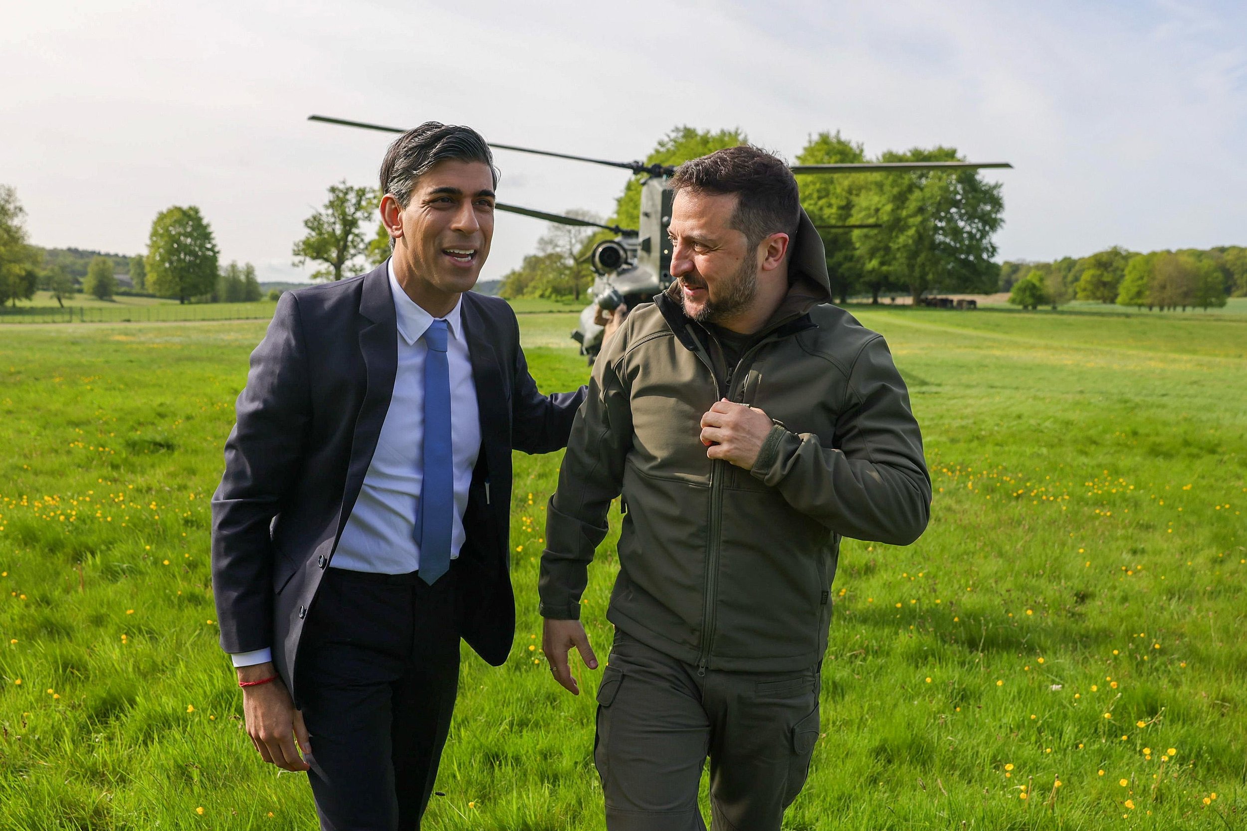 Two men are having a conversation outdoors in a grassy field with trees in the background. One man is wearing a suit and tie, and the other is dressed in casual outdoor clothing. There is a helicopter behind them.