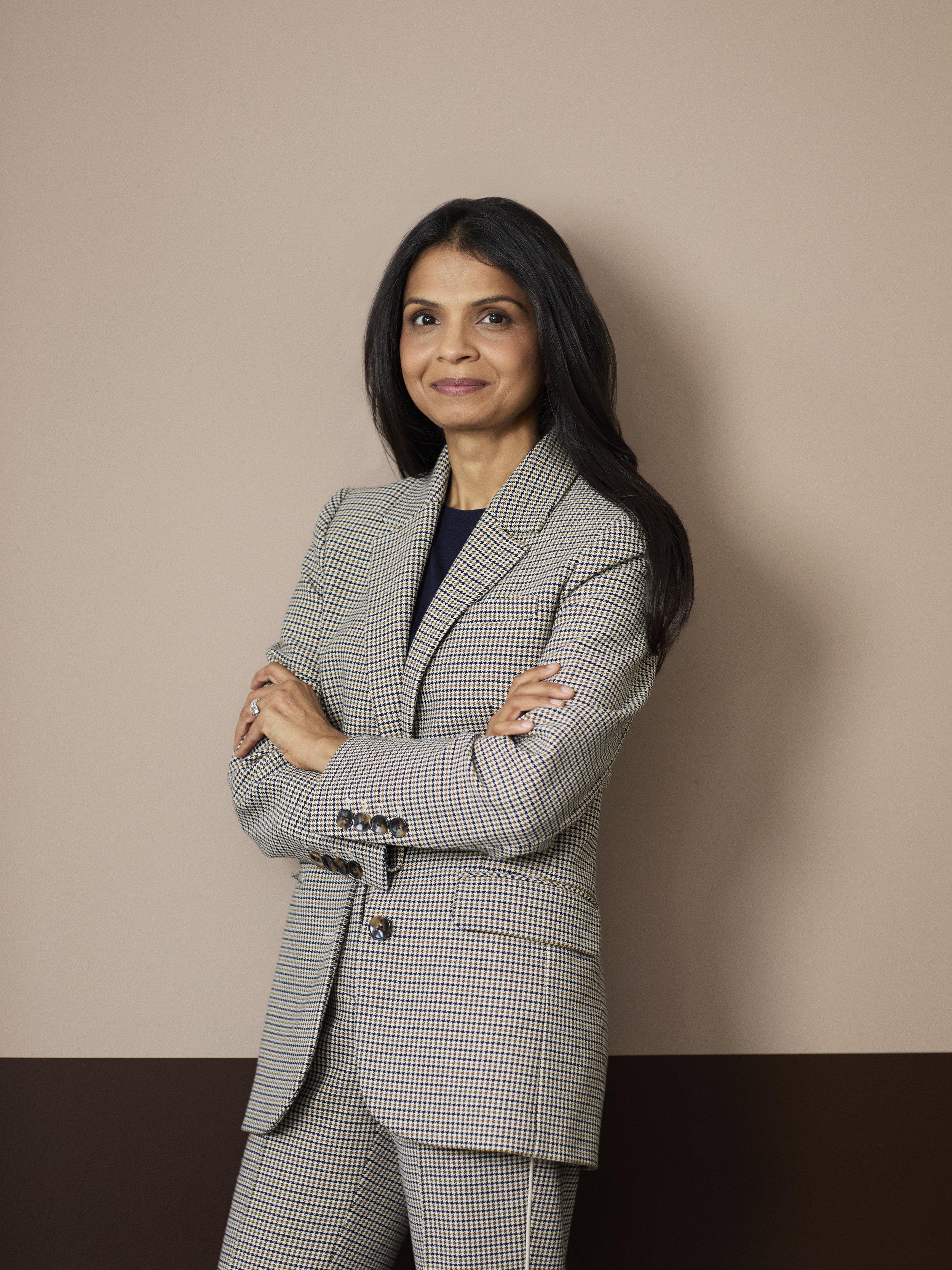A woman with long black hair, wearing a checkered blazer with rolled-up sleeves, standing with arms crossed against a plain beige background.