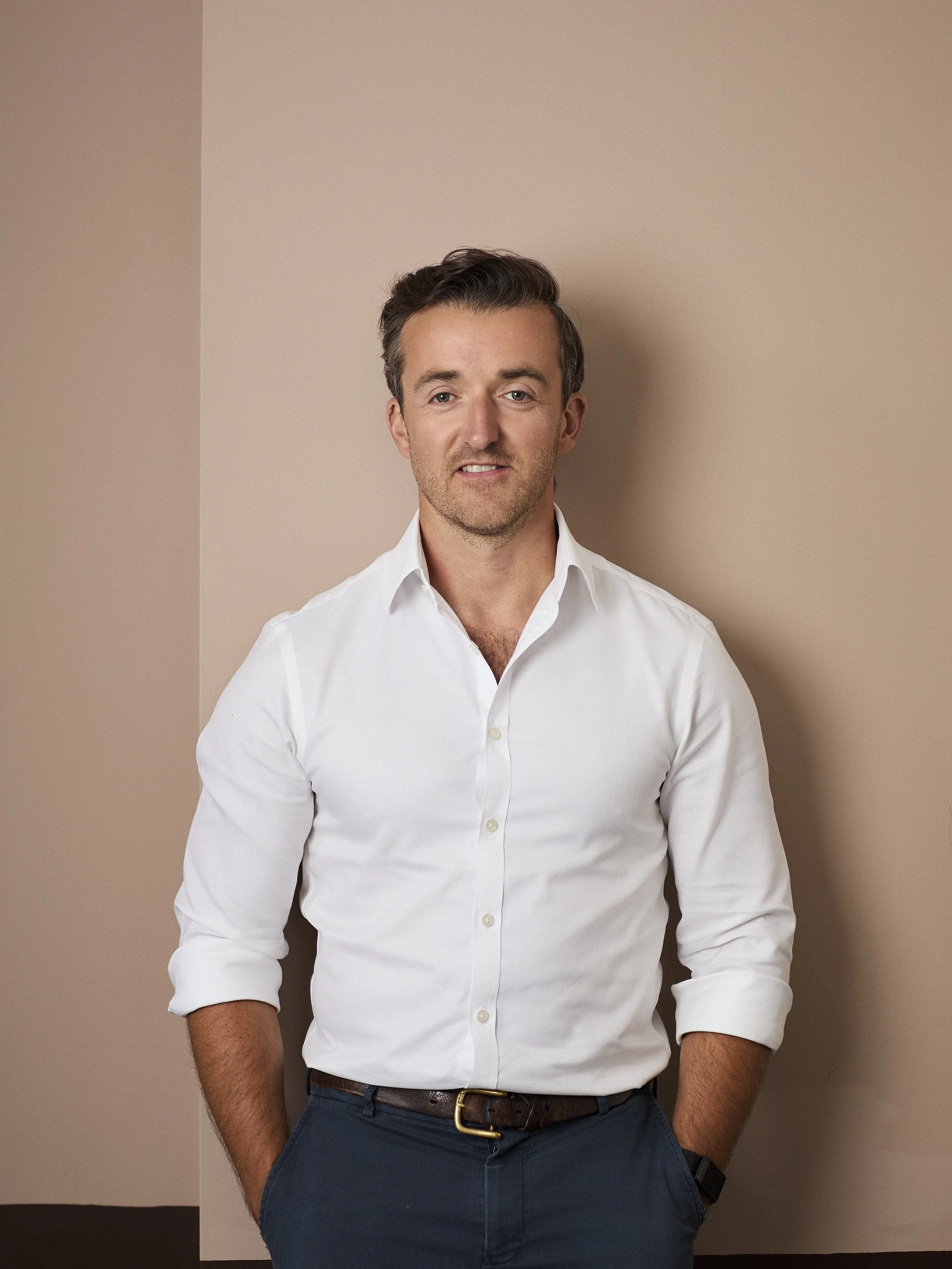 A man with short dark hair, wearing a white dress shirt with rolled-up sleeves, dark blue pants, and a black belt, standing against a plain beige and dark background.