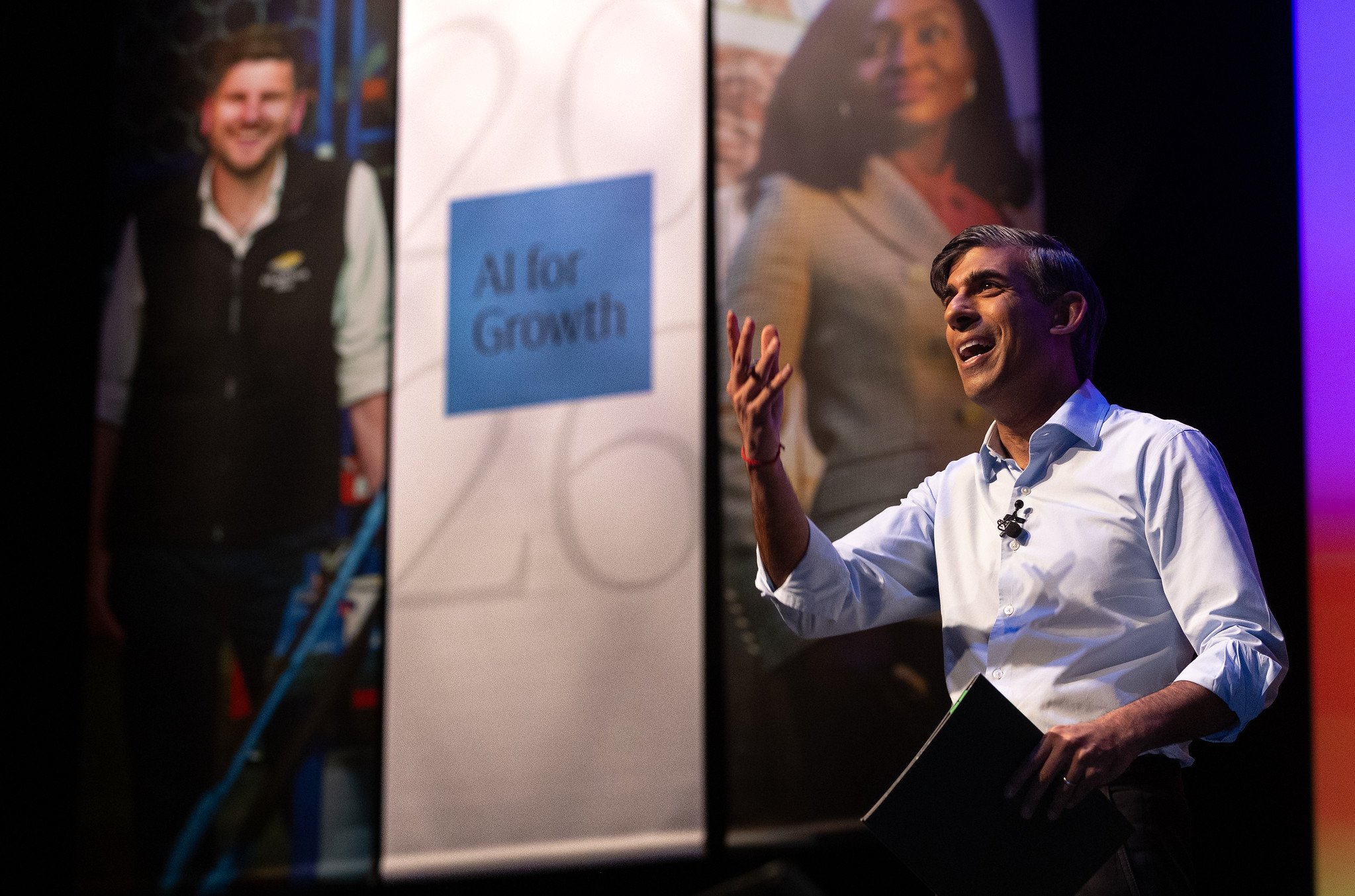 A man in a white shirt appears to be speaking or presenting, with a smile and expressive hand gestures. In the background, there are large posters featuring diverse individuals, one man and one woman. The setting looks like a conference or seminar, with dark lighting and colorful side panels.