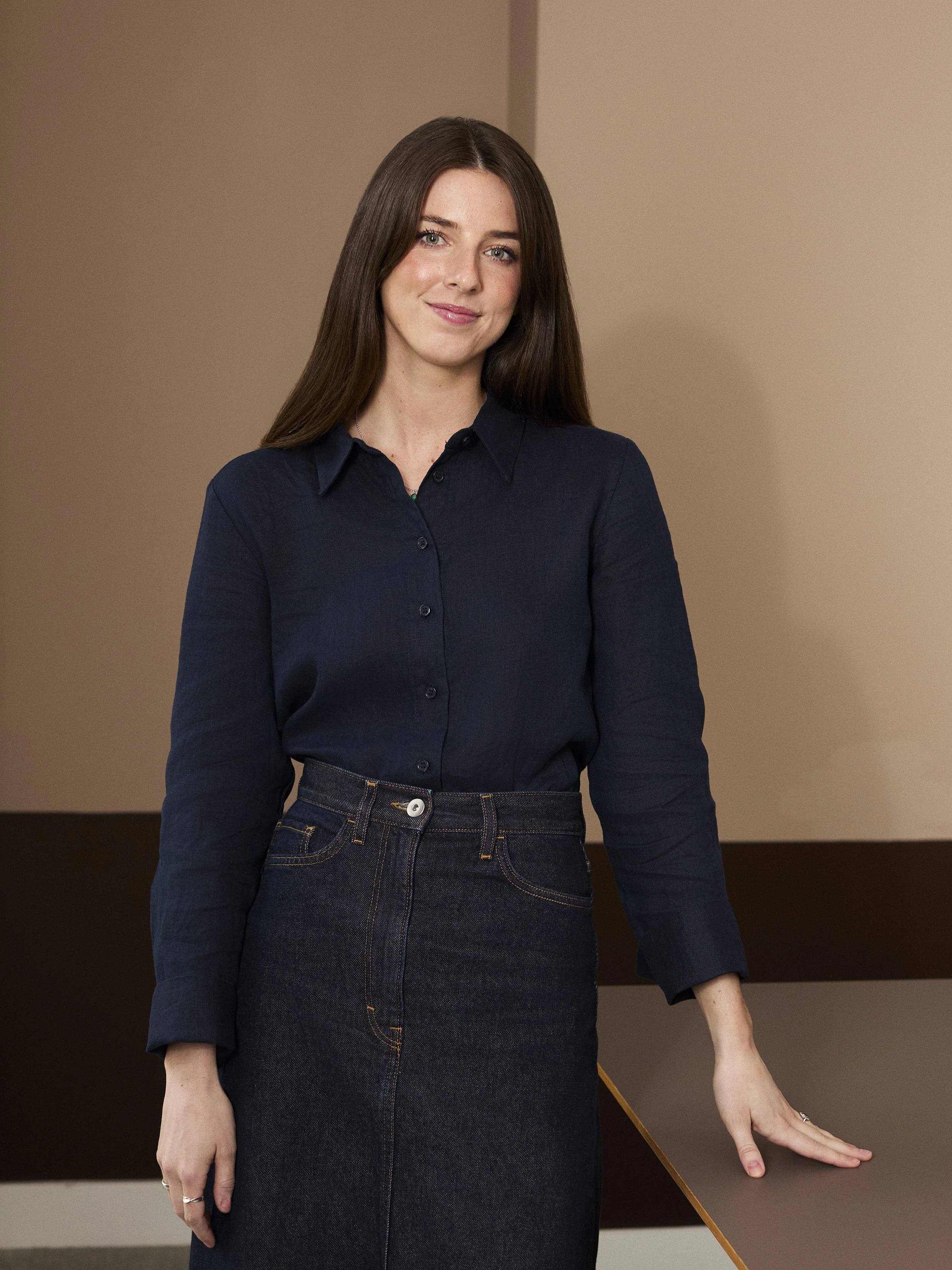 A woman with long brown hair, wearing a dark blue button-up shirt and a black denim skirt, standing indoors against a beige wall and a dark brown table.