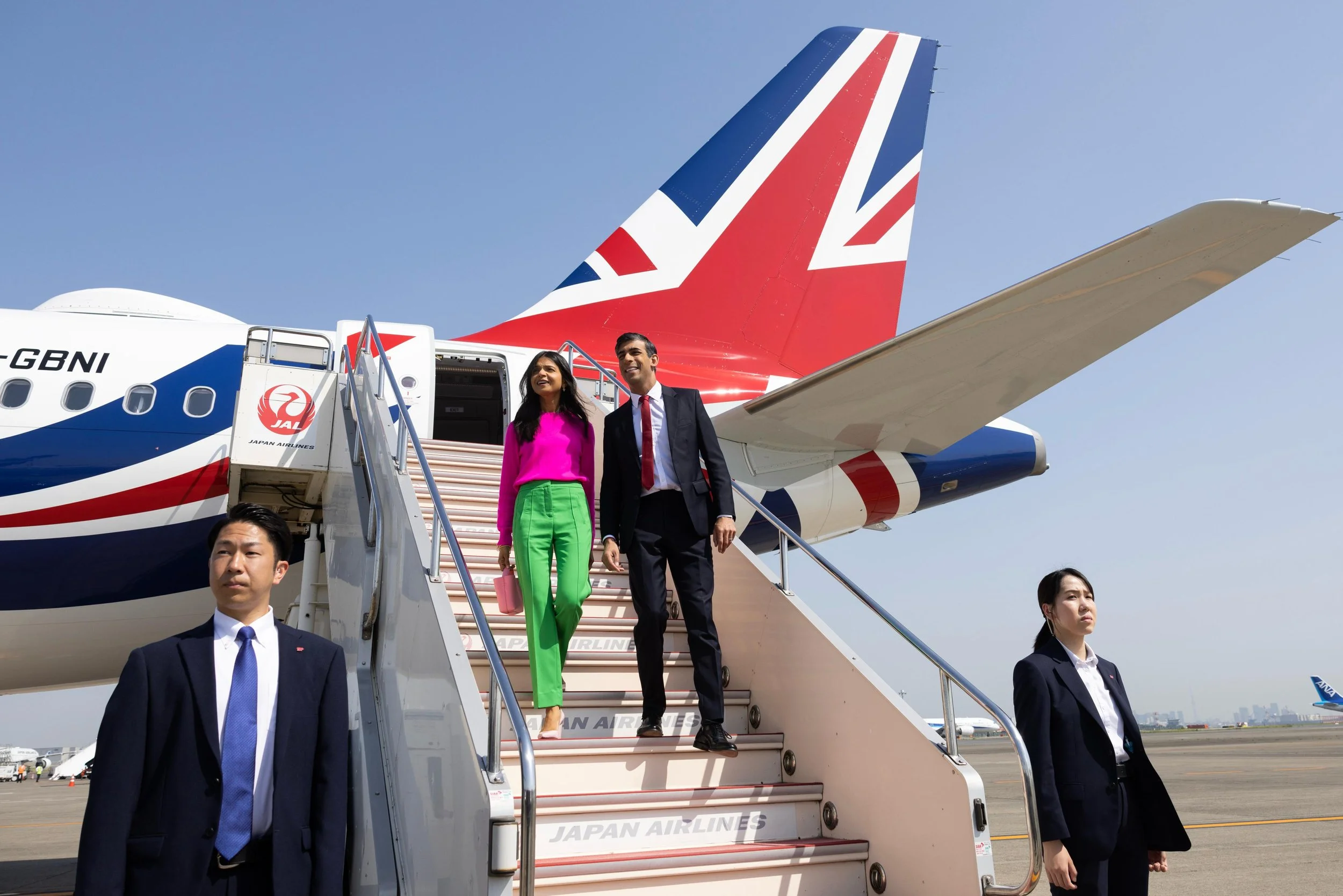People disembarking an airplane with Japan Airlines logo, ascending stairs, and security personnel nearby, at an airport under a clear sky.