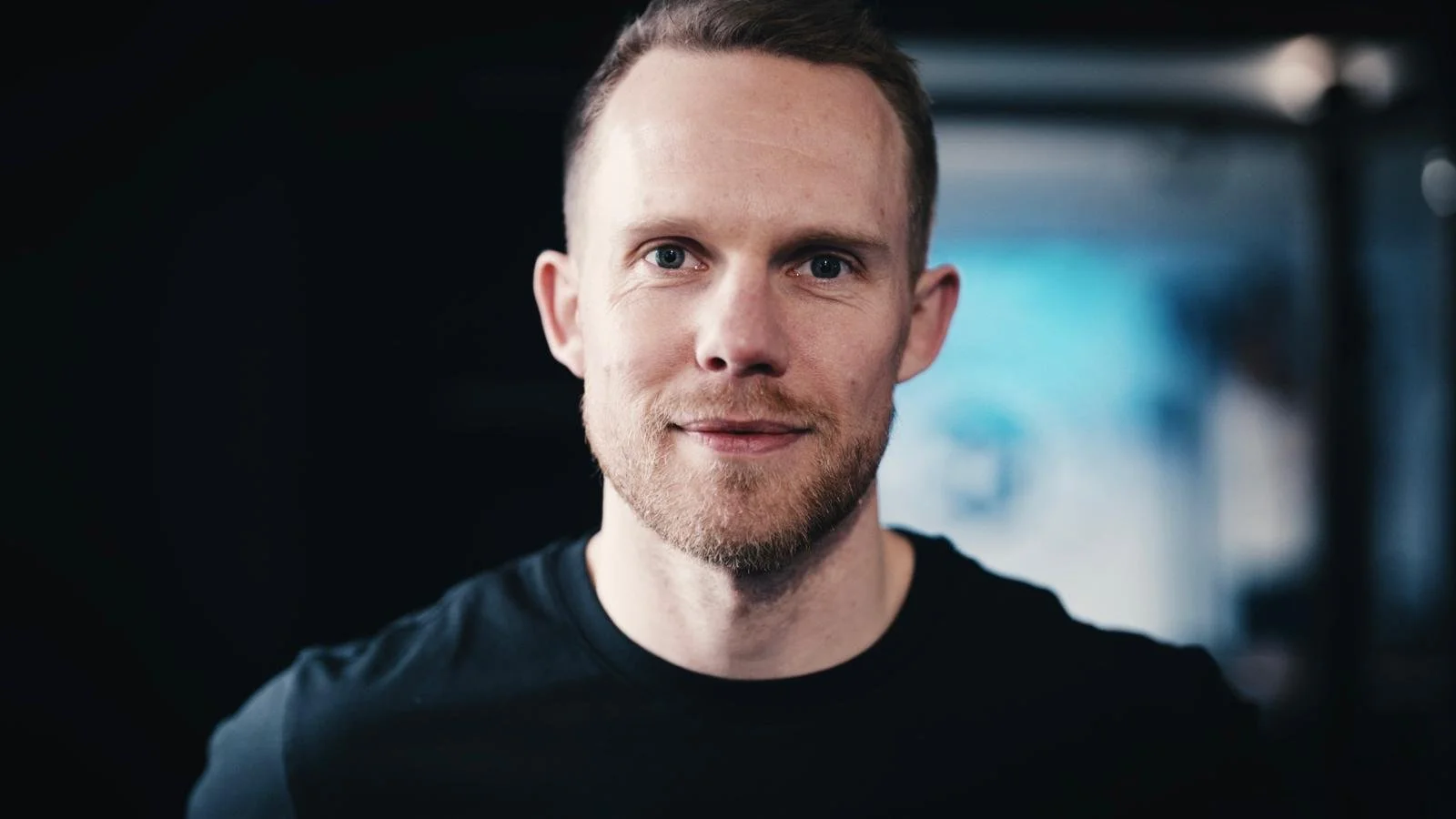 A man with light brown hair, beard, and blue eyes in a close-up portrait, wearing a black shirt, smiling slightly, with a blurred background.