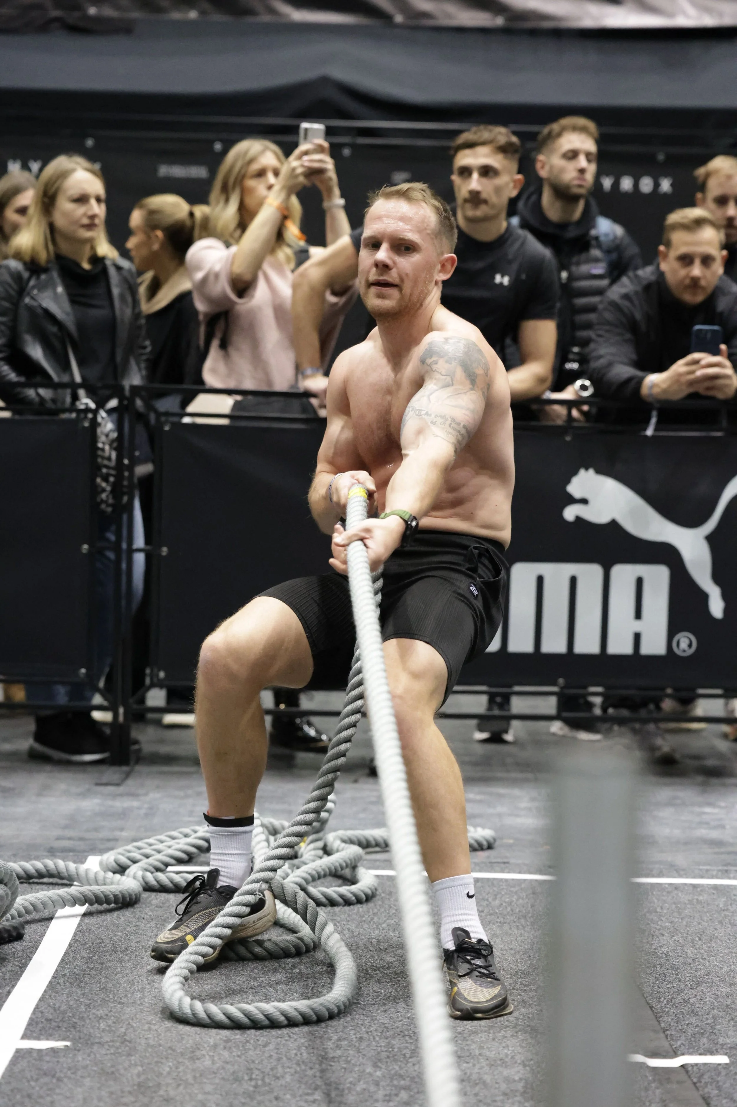 A shirtless man using battle ropes for exercise in a gym, with a group of people watching and taking photos in the background. Competing in hyrox