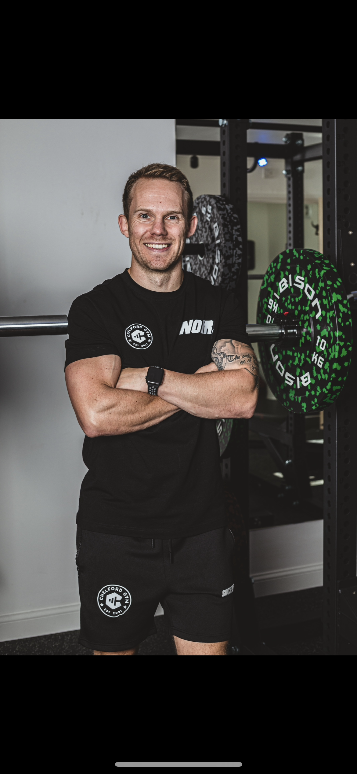 A muscular man standing in a gym, smiling with arms crossed, wearing a black workout shirt and shorts with a gym logo, next to weightlifting equipment.
