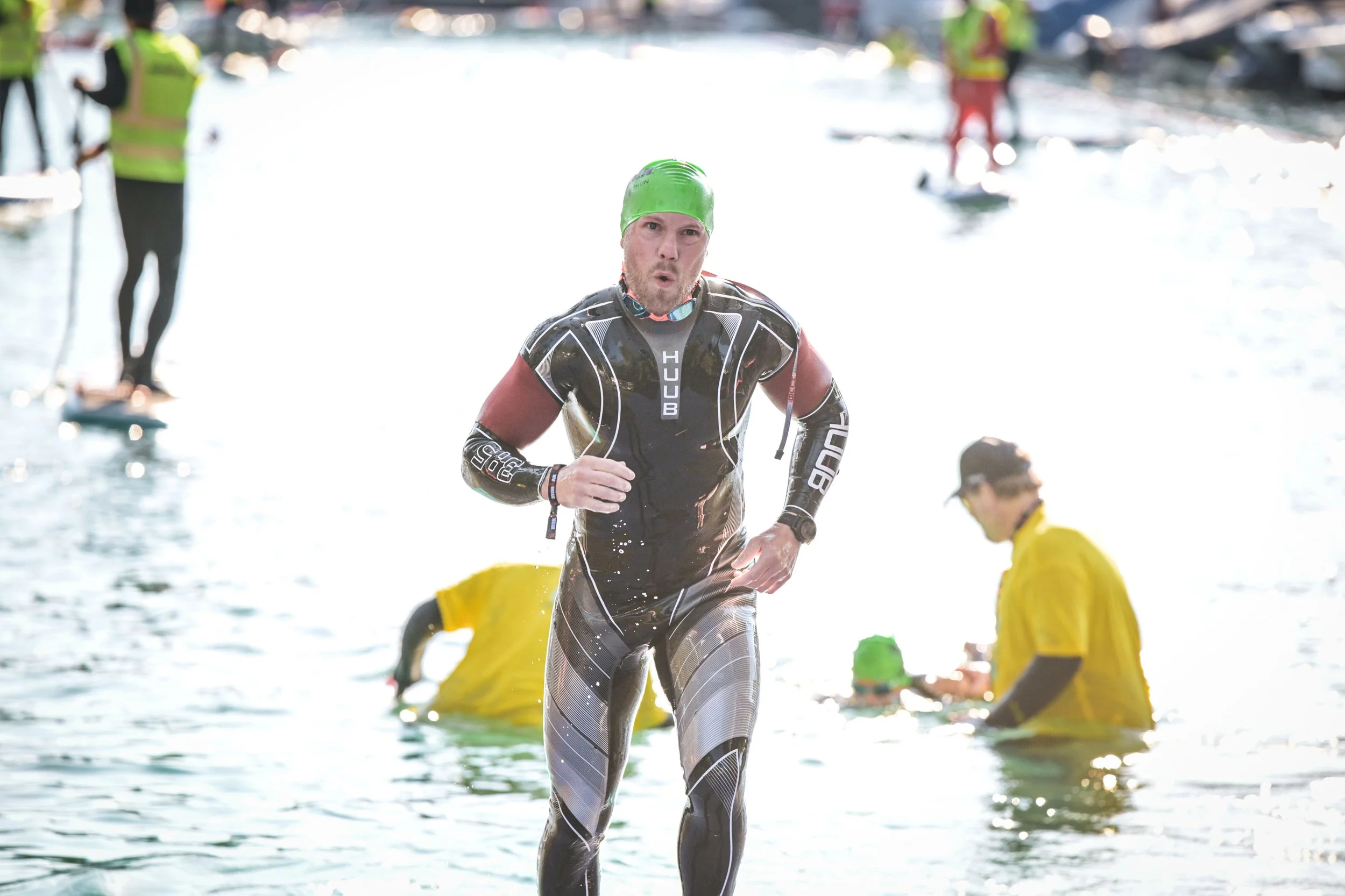 Male athlete in a wetsuit and green cap running through water during a triathlon with spectators and other participants in the background.