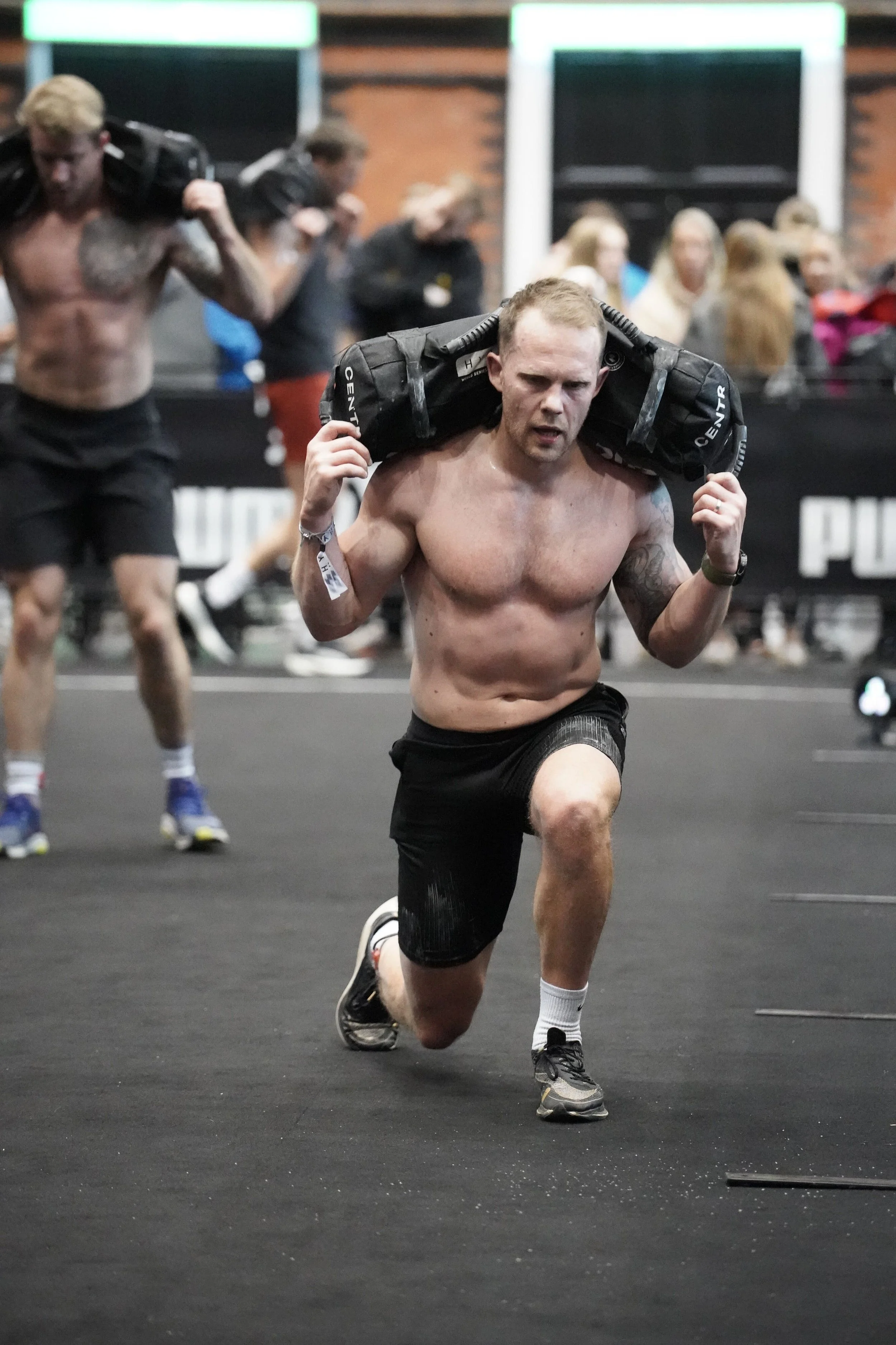 A shirtless man doing walking lunges during a hyrox competition.
