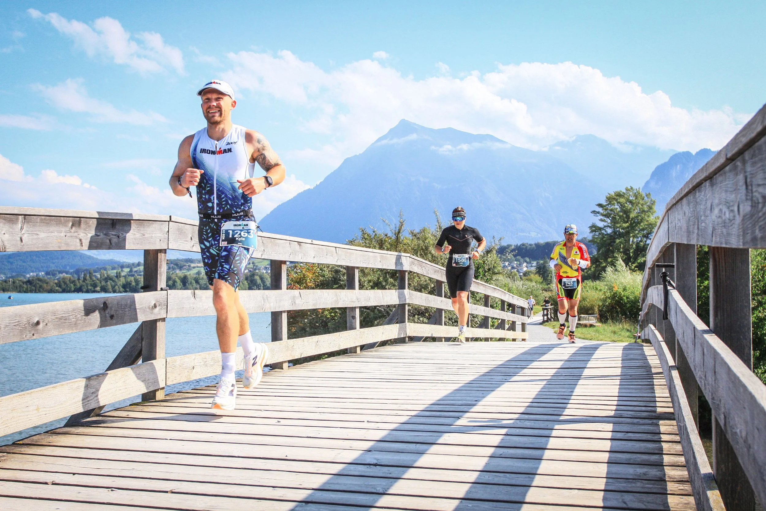 Three runners participating in a race on a wooden bridge over water, with mountains and blue sky in the background. Man competing in Ironman Switzerland.