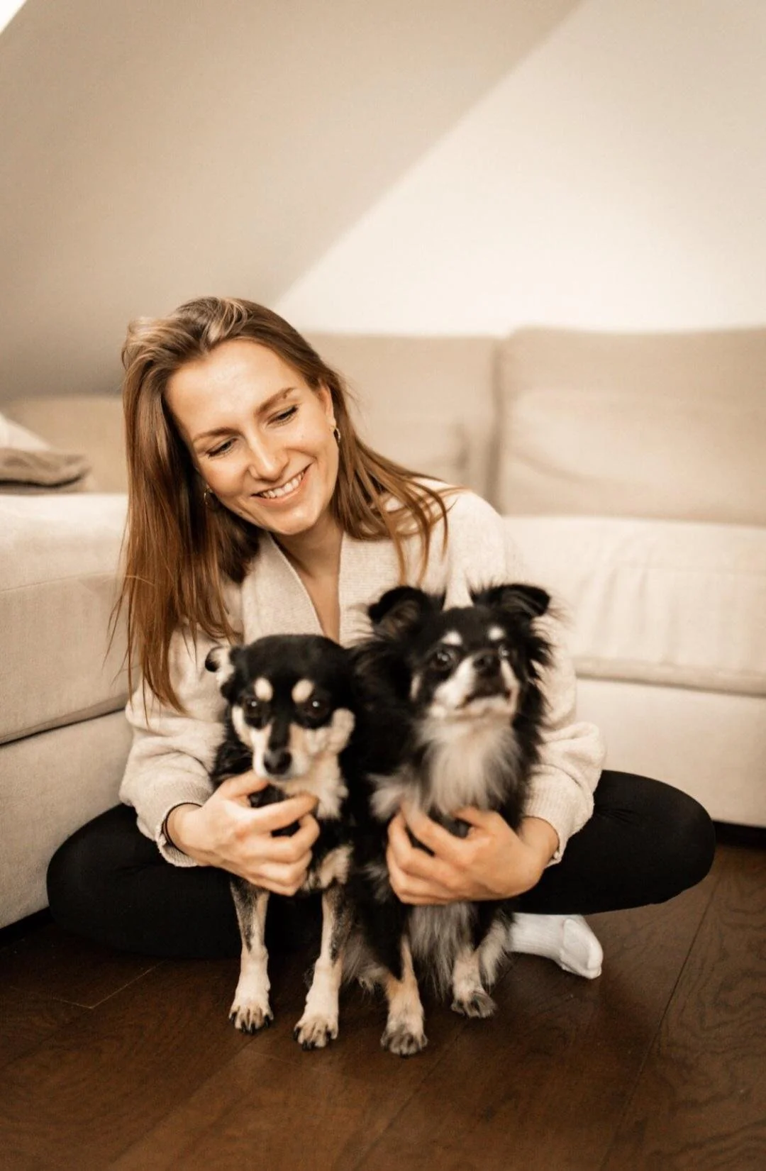 Eine lachende Frau hält zwei kleine Hunde, sitzend auf dem Boden in einem Wohnzimmer.