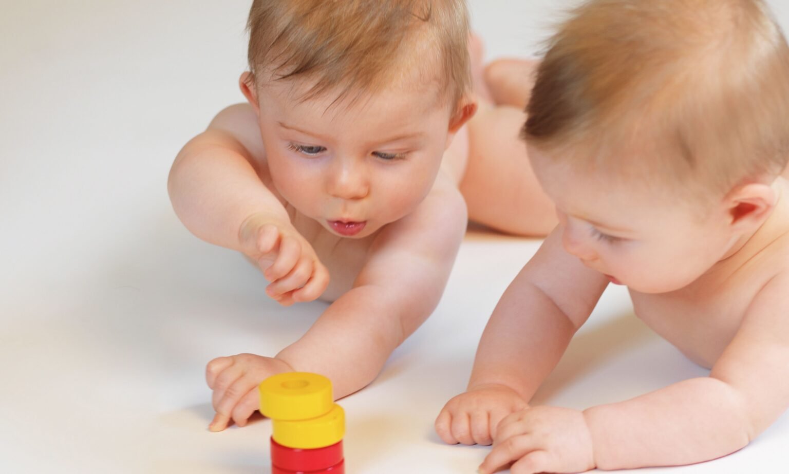Zwei nackte Babys liegen auf dem Bauch und spielen mit bunten Spielsteinen, einem roten und zwei gelben.