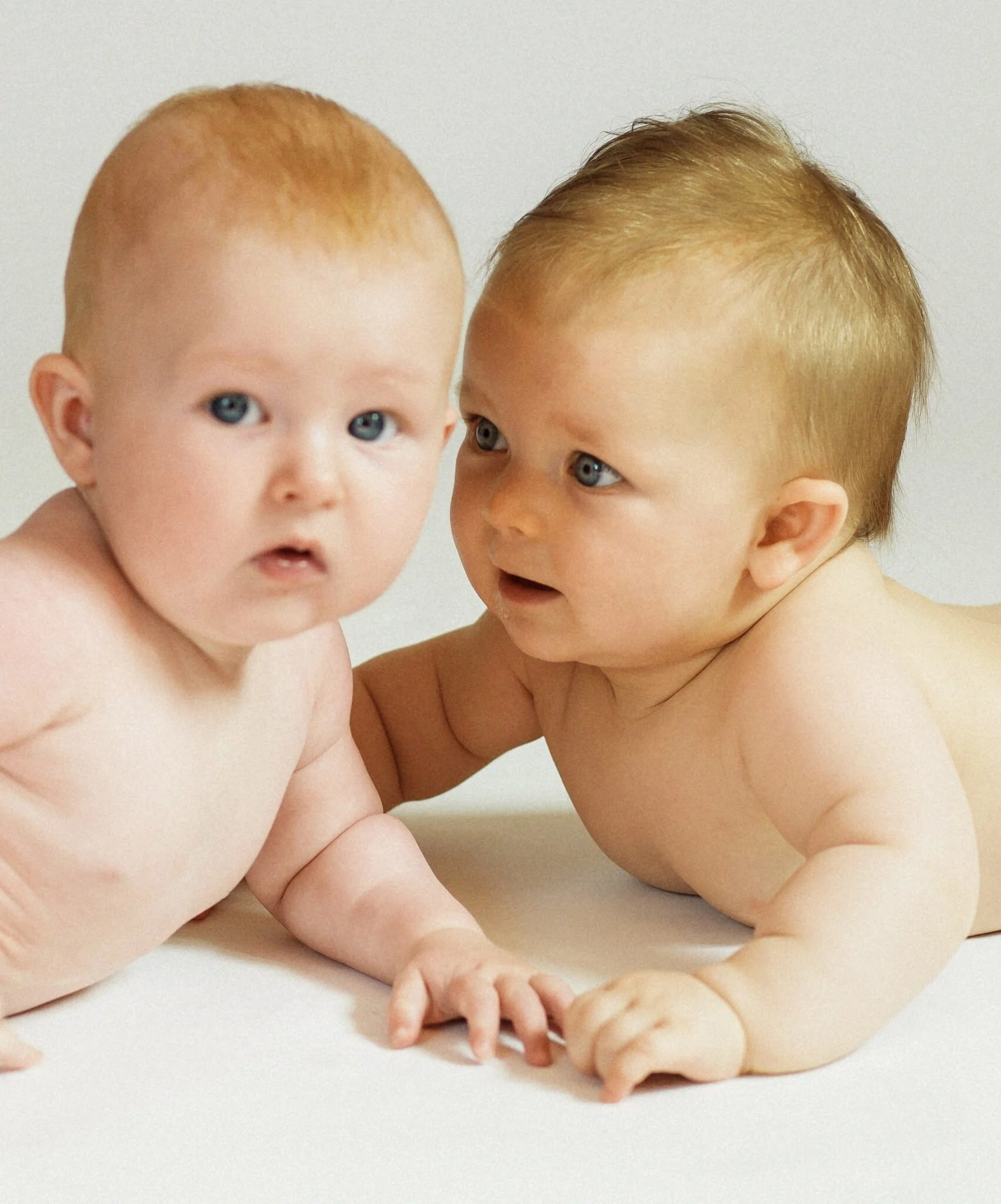 Zwei nackte Babys, die auf einem weißen Untergrund liegen, schauen aufmerksam und neugierig.