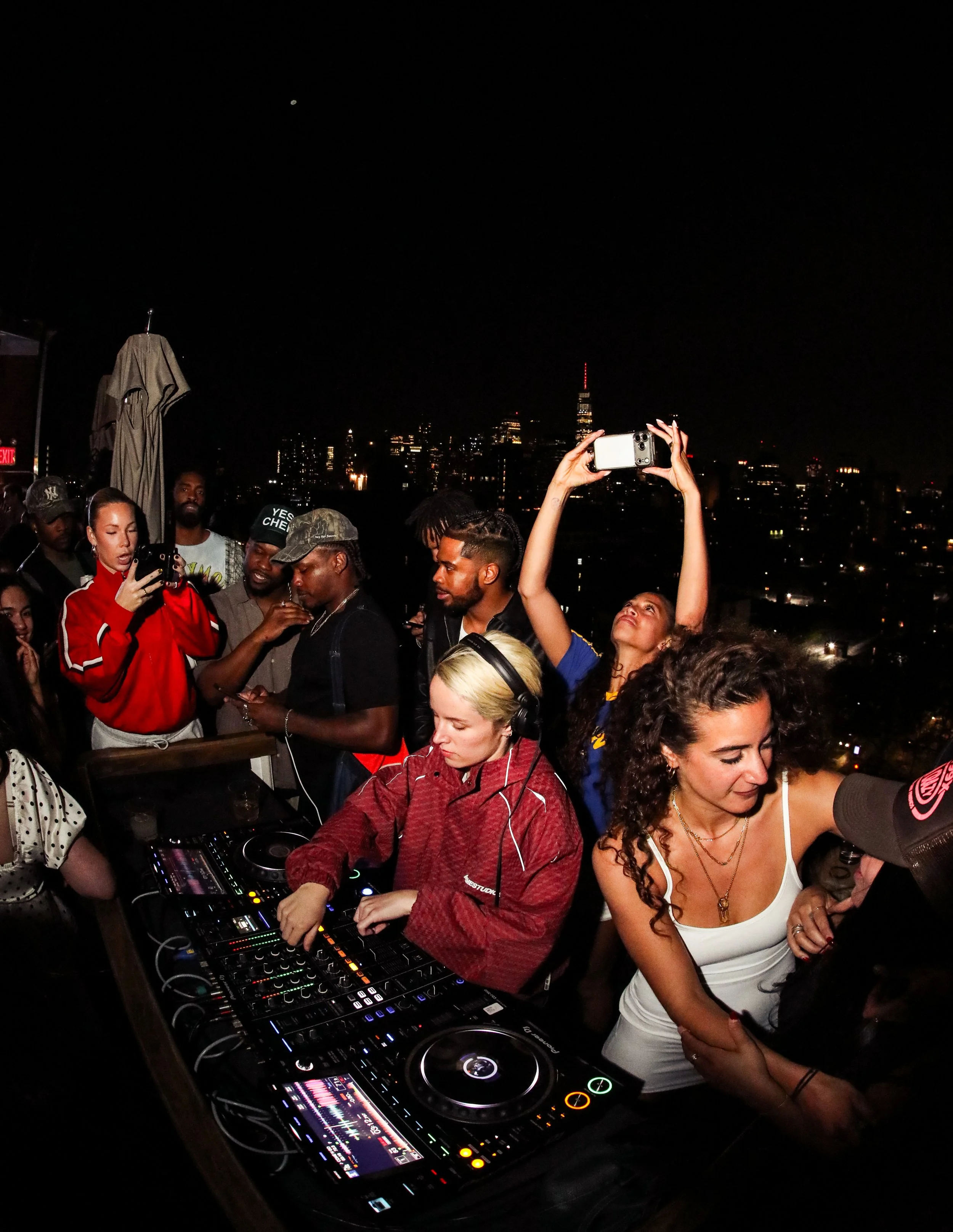 People dancing and socializing on a rooftop at night, with city skyline in the background, including tall buildings and a lit-up tower, while a DJ mixes music on a turntable.