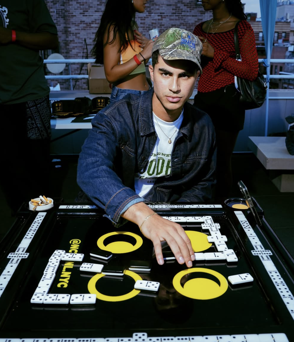 Young man playing dominoes at a table during a social gathering, with people standing behind him in a festive outdoor setting.