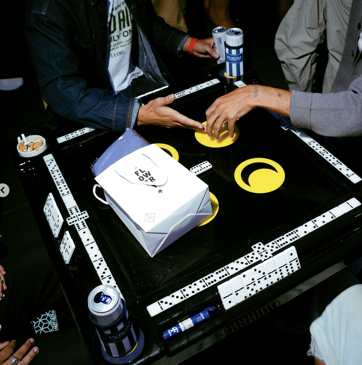 People playing dominoes around a table with domino tiles, drinks, and a shopping bag on it, with some cigarette butts in an ashtray.