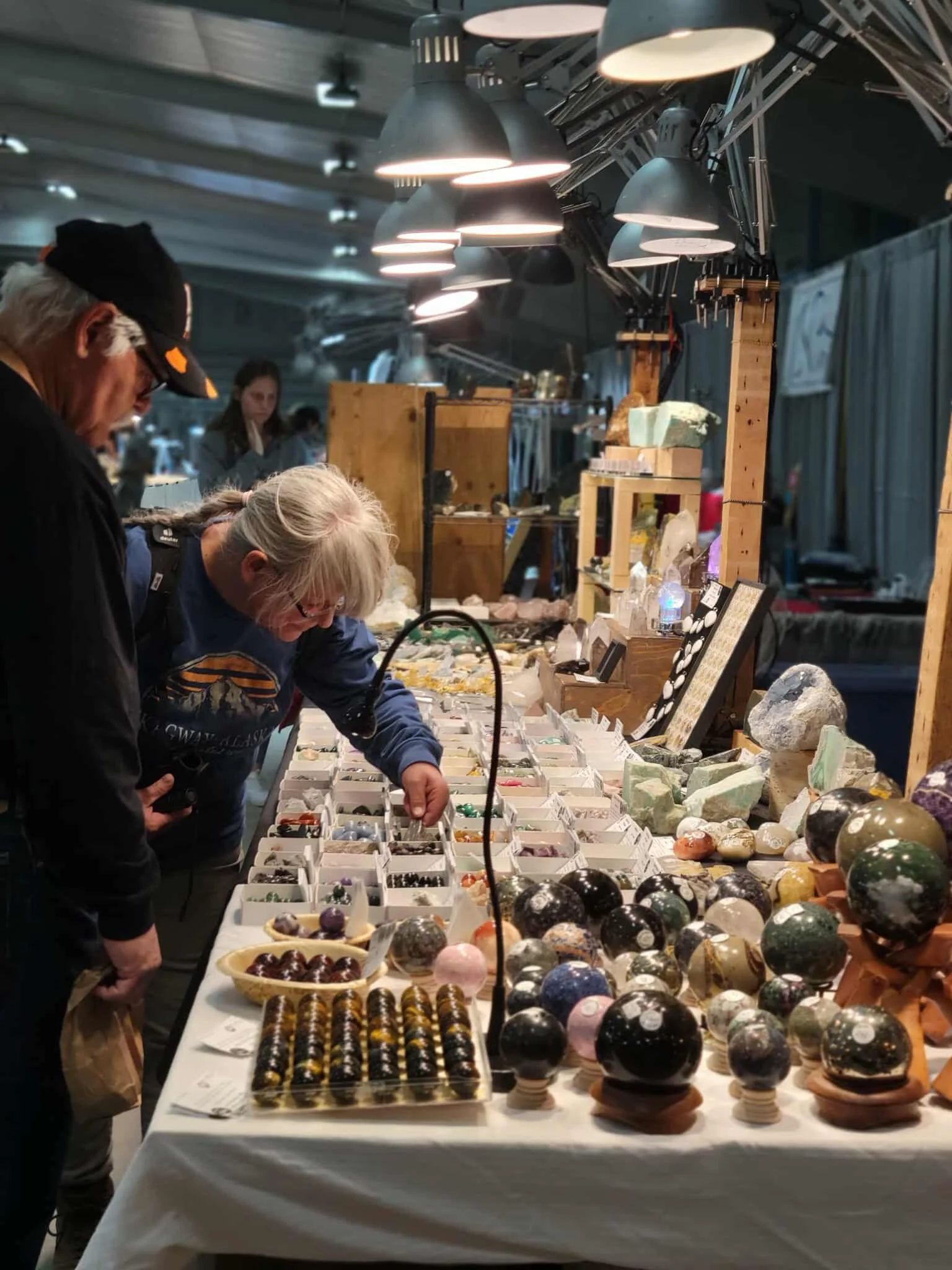 People examining a display of colorful polished gemstones and mineral specimens at an indoor market or exhibition with overhead lighting.