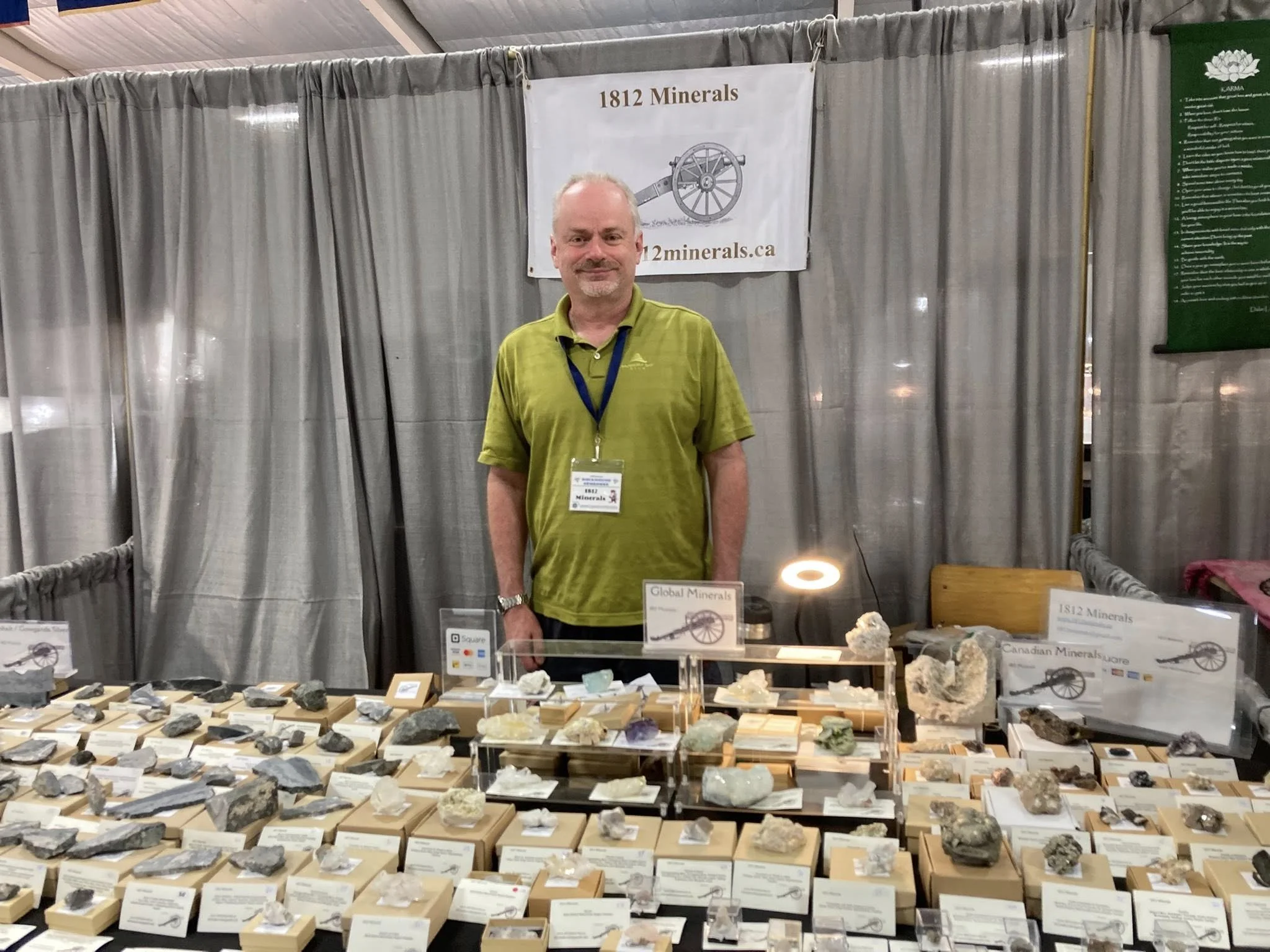 A man wearing a green shirt and lanyard standing behind a display of various mineral specimens at a mineral show. Behind him is a banner that reads '1812 Minerals' and a website URL. The display table has rocks and minerals arranged on small stands a