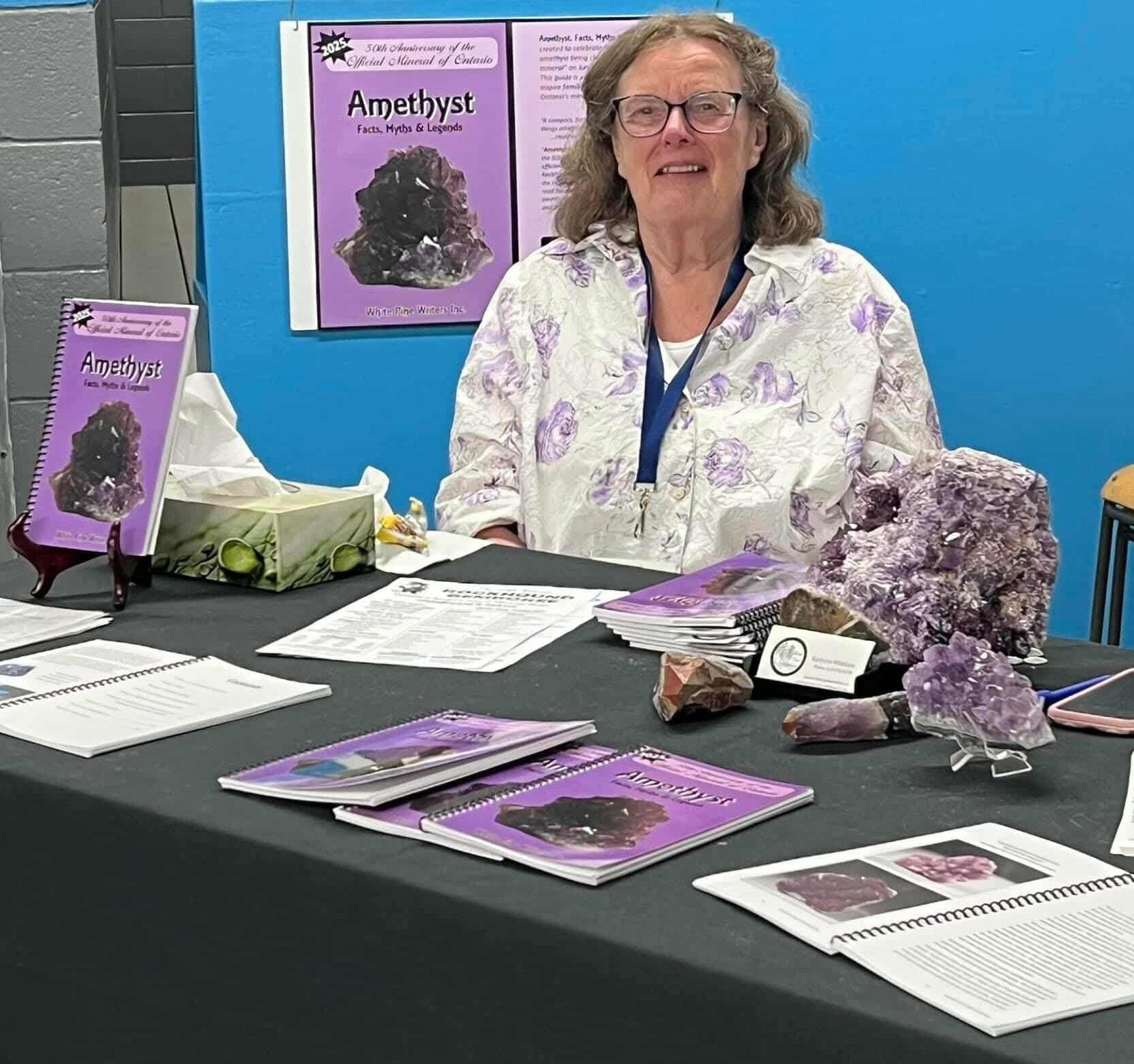 A woman sits behind a table at a display booth about amethyst crystals, with informational pamphlets, rocks, and a small framed sign on the table, and a large poster about amethyst on the wall behind her.