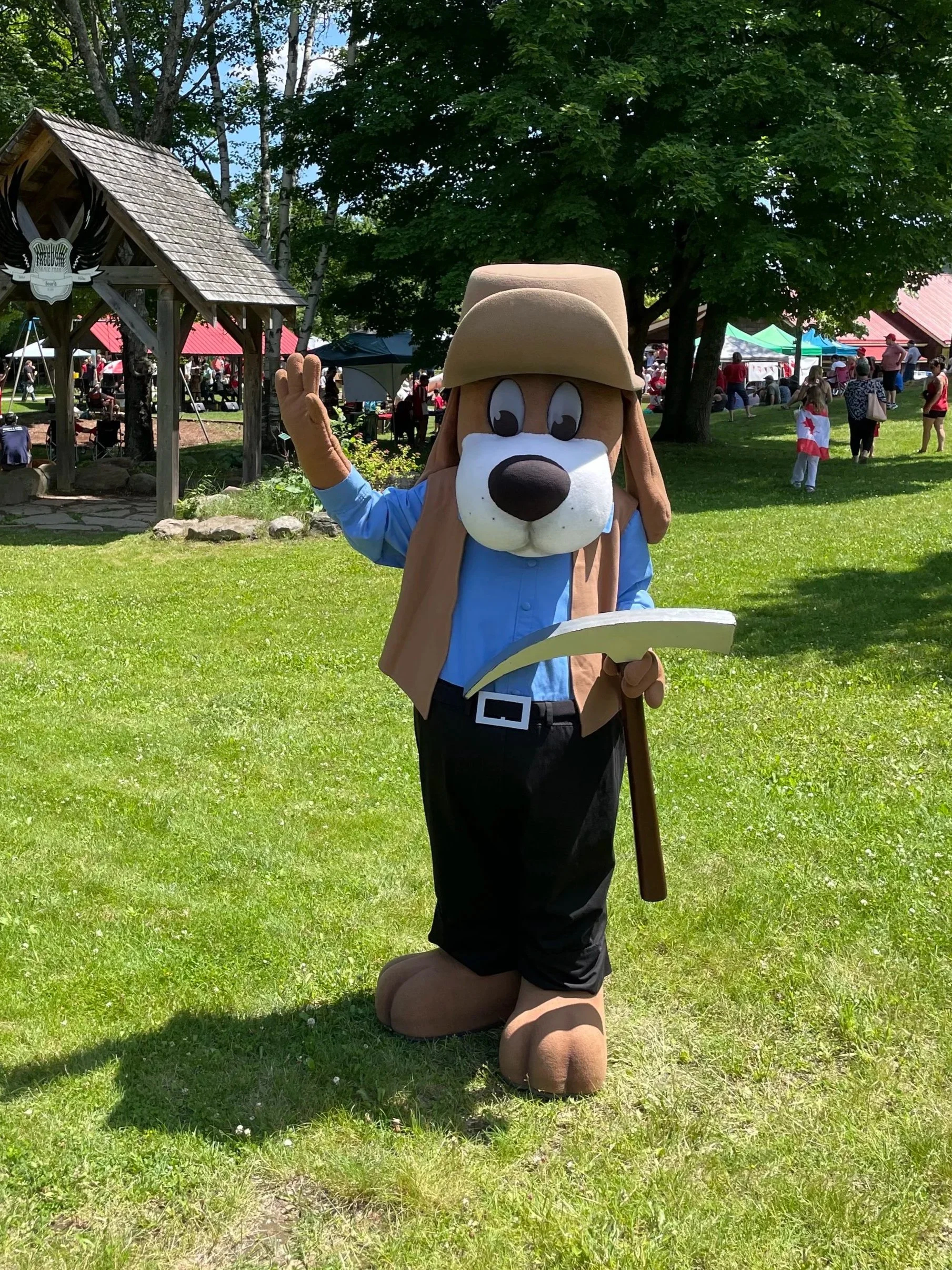 A person dressed in a dog costume, kneeling on grass at an outdoor event with tents and people in the background.