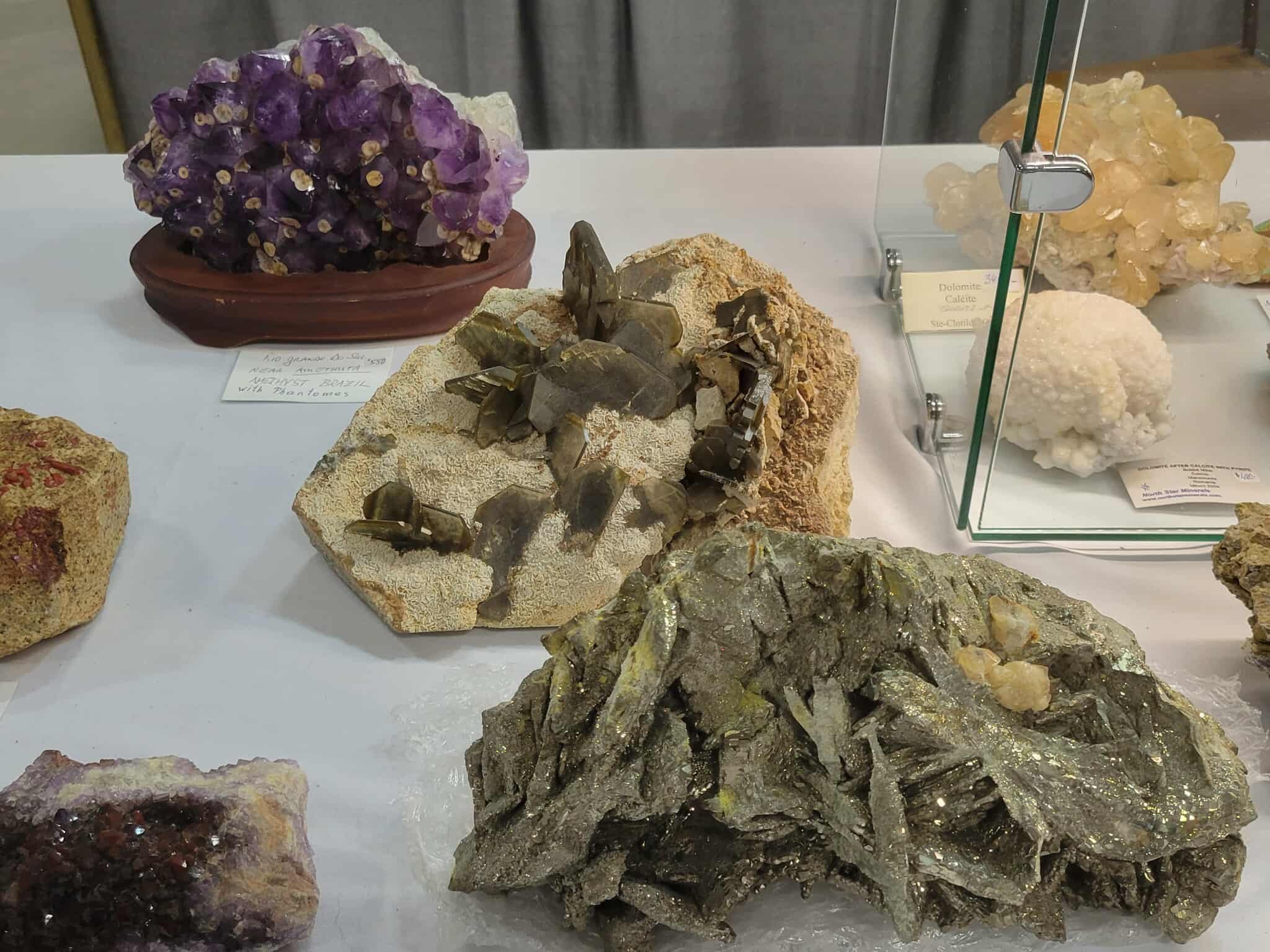 Various mineral and crystal specimens displayed in a glass case, including a purple crystal cluster on a pot, metallic and earthy mineral rocks, and other mineral formations.