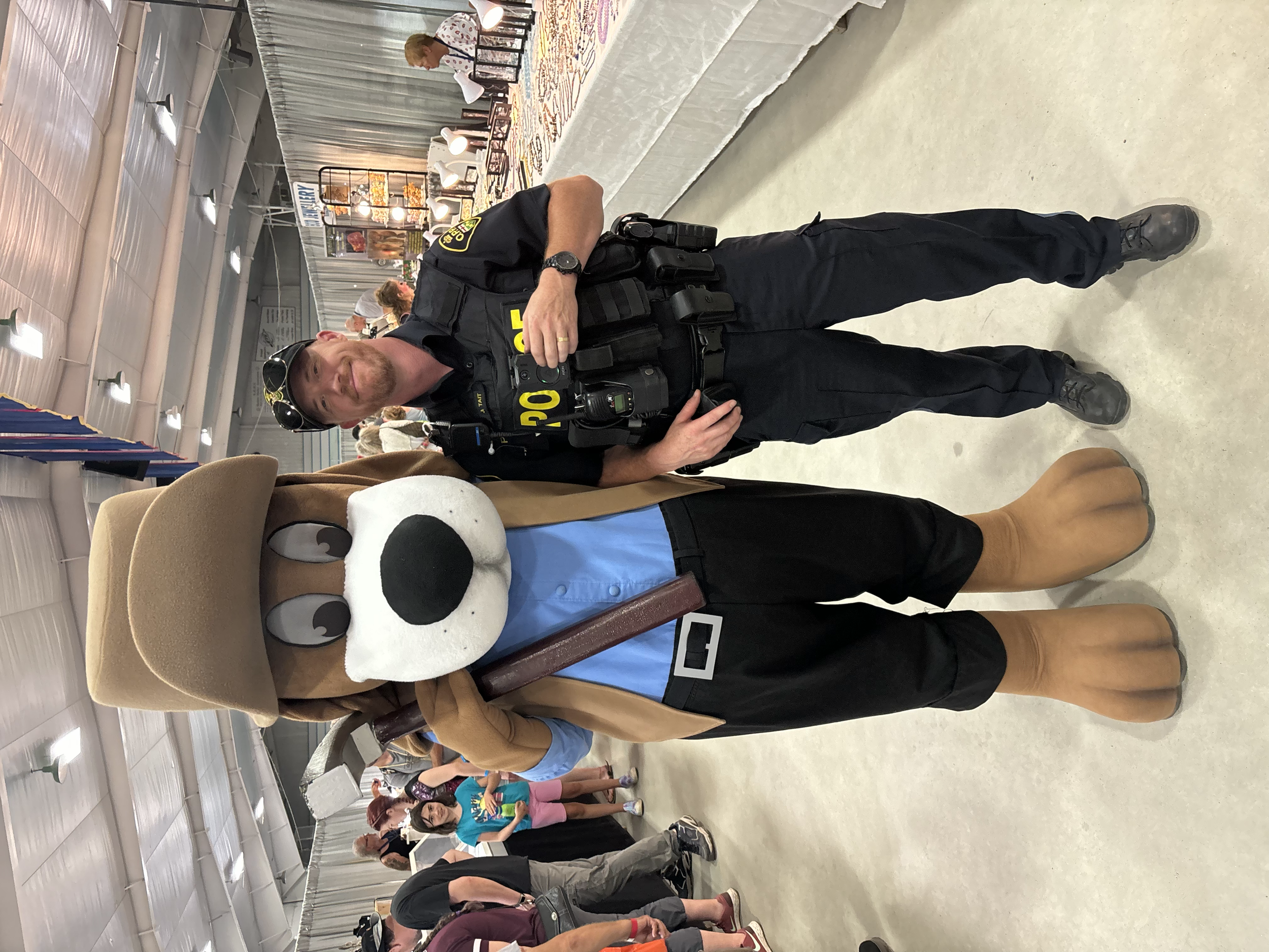 A police officer and a person in a large dog costume with a tan hat and brown shorts at an indoor event. The officer is wearing a uniform with a badge, radio, and a belt with various equipment. The person in the costume appears to have their arm arou