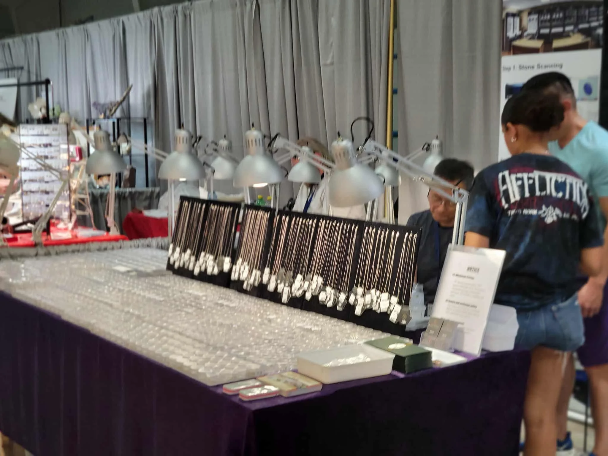 Store display table with jewelry or small decorative items under bright lamps, with people browsing and a curtain backdrop.