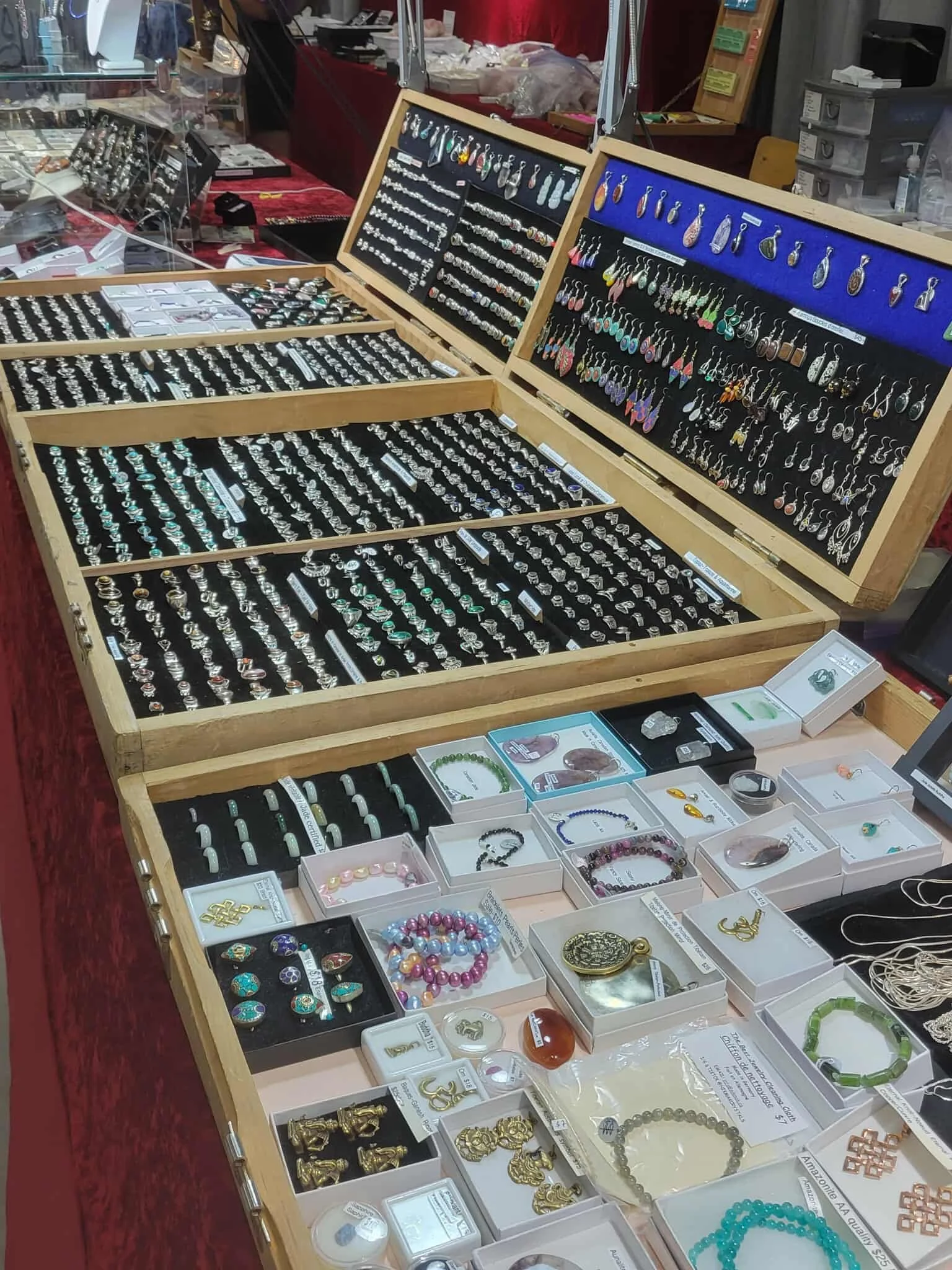 Display of various jewelry items including rings, earrings, bracelets, and pendants arranged on wooden cases and in small boxes at a market stall.