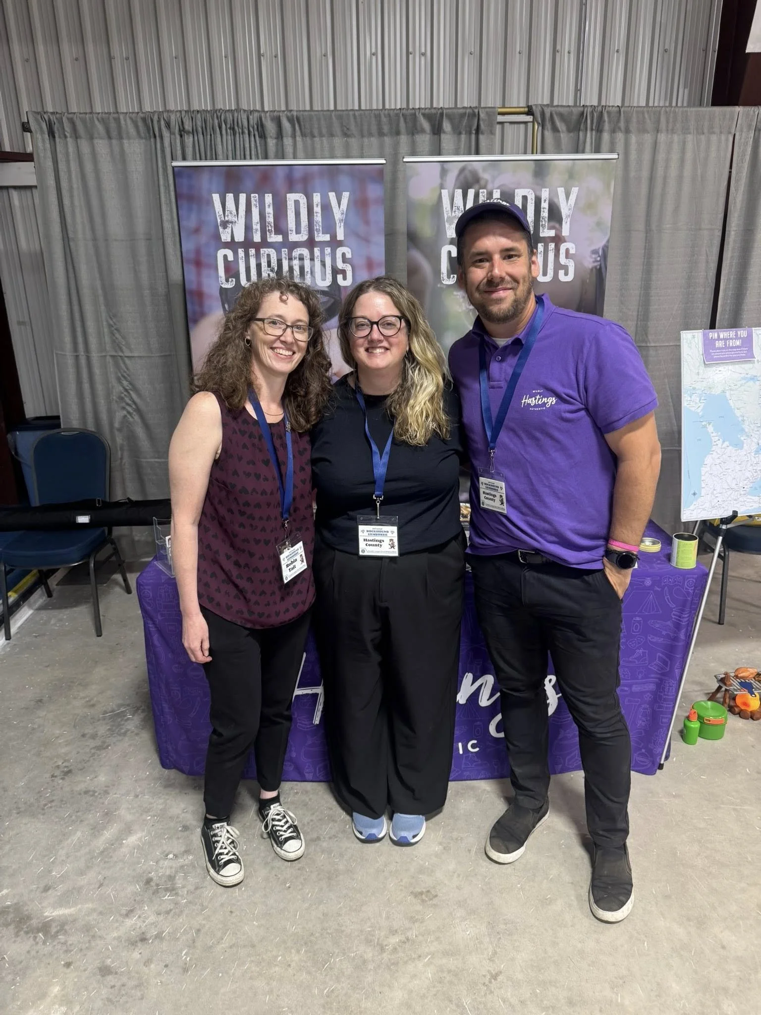 Three women standing together at an indoor event with a backdrop that reads "Wildly Curious." They are smiling and wearing name tags, with a table partially visible in front of them and children’s toys on the floor nearby.