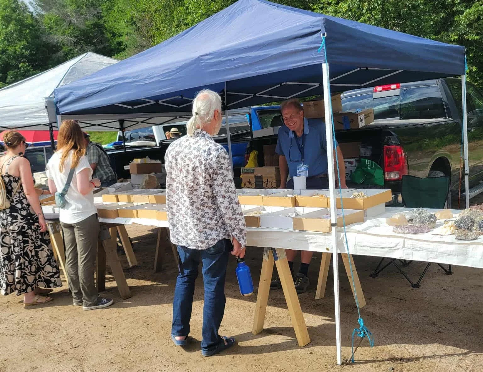 People shopping at an outdoor market stall under a blue canopy tent, with a man behind the stall and customers looking at rocks or minerals on display.