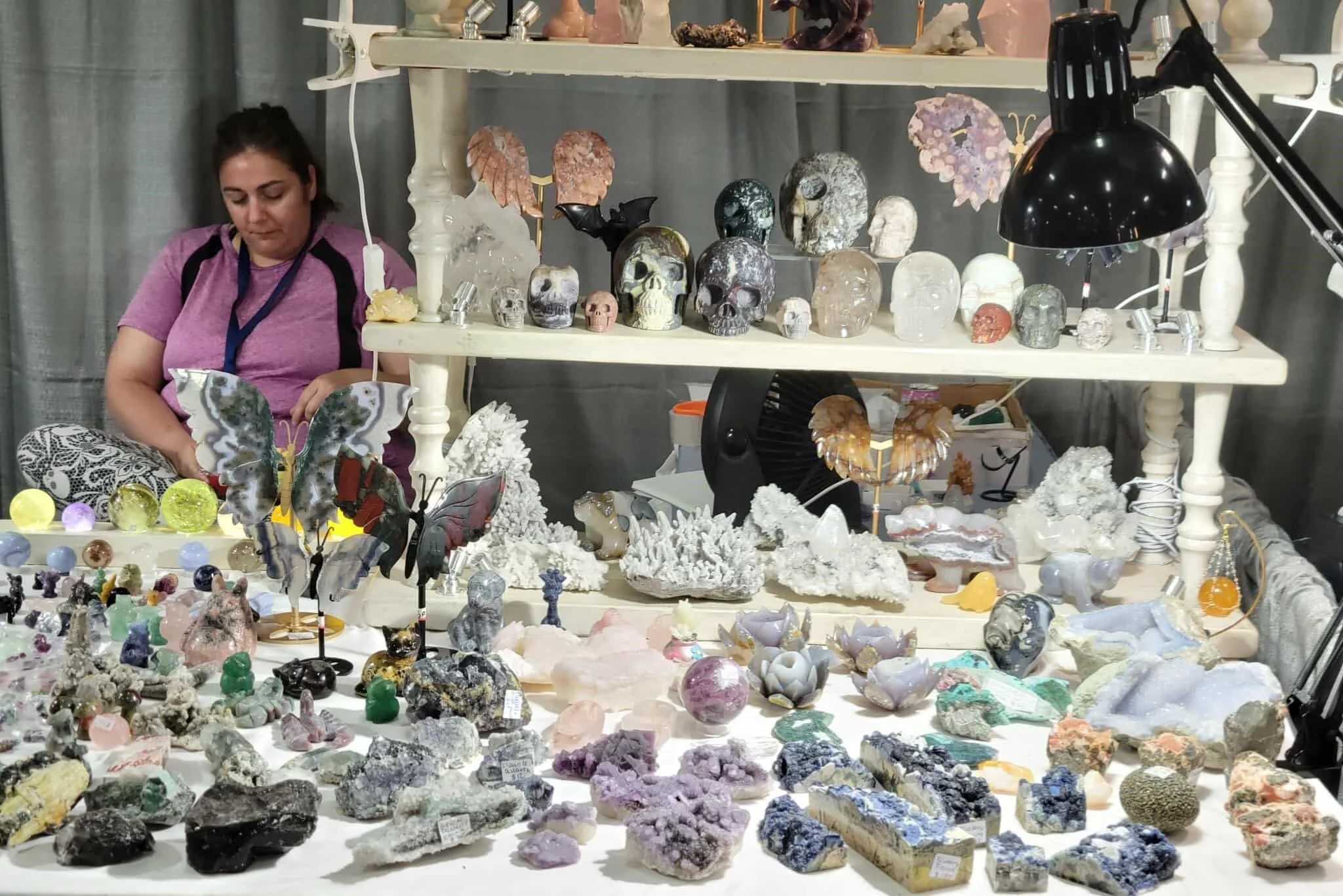A display of various minerals, crystals, and gemstones on tables and shelves at a market or exhibit, with a woman in purple and black clothing inspecting the items.