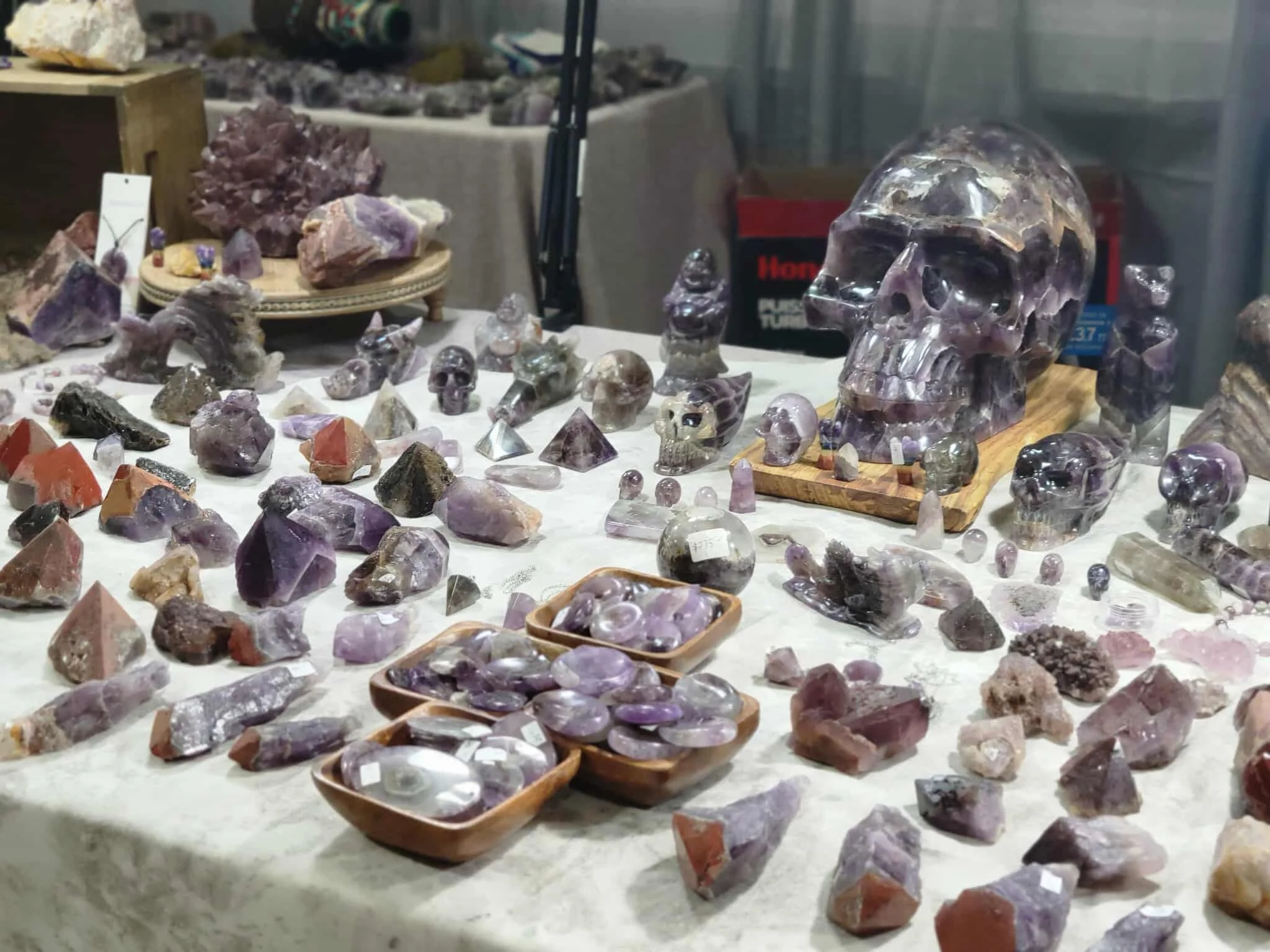 Display of various mineral and crystal specimens, including a carved skull made from purple and clear crystals, small skull figurines, and geodes on a table at an indoor mineral show or shop.