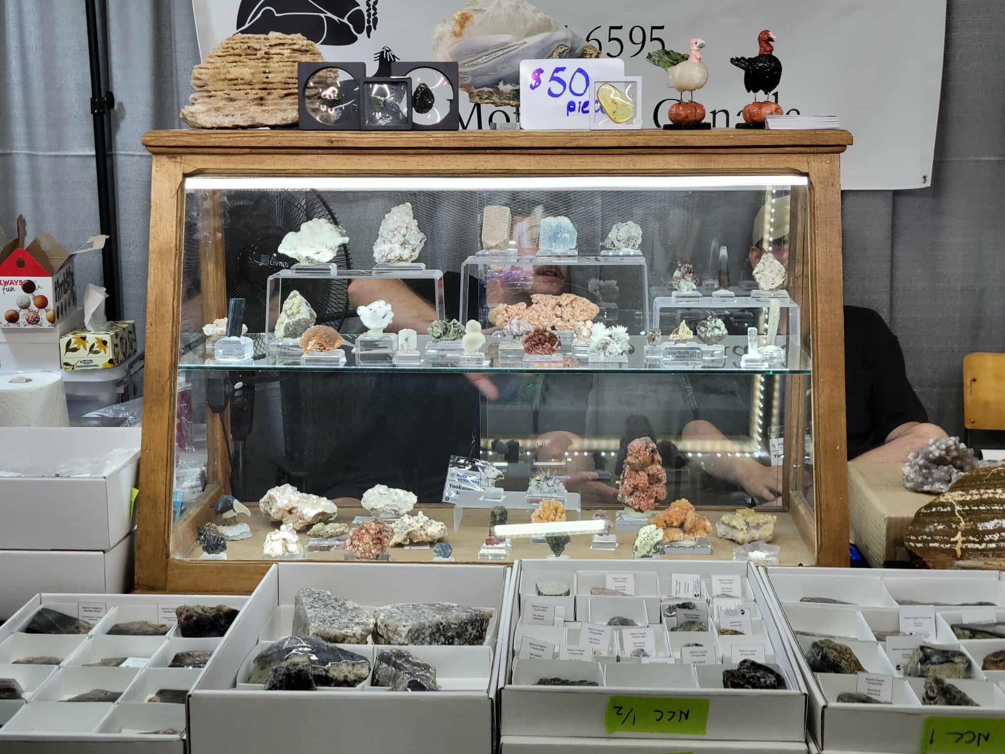 Display case filled with various mineral and rock specimens. On top of the case, there are decorative items and a sign indicating prices. The background features a display advertisement or sign.