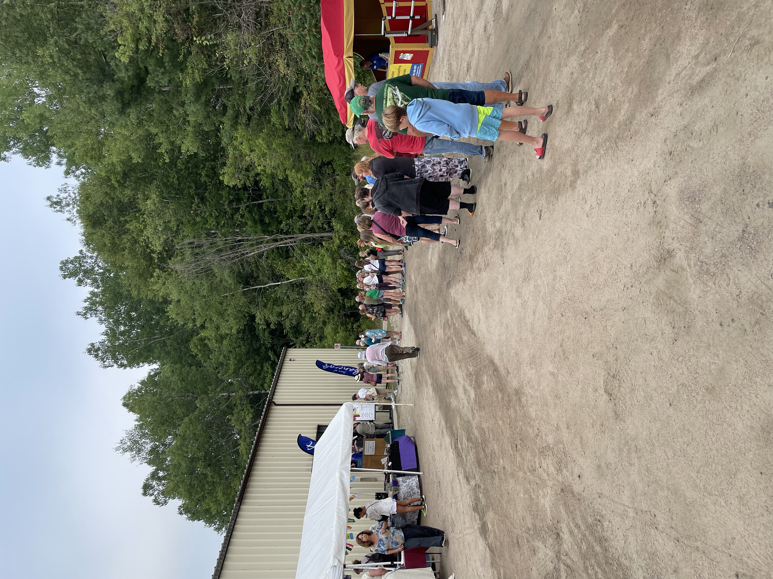 People standing in line outside at a community event under trees, with a tent and booths set up.