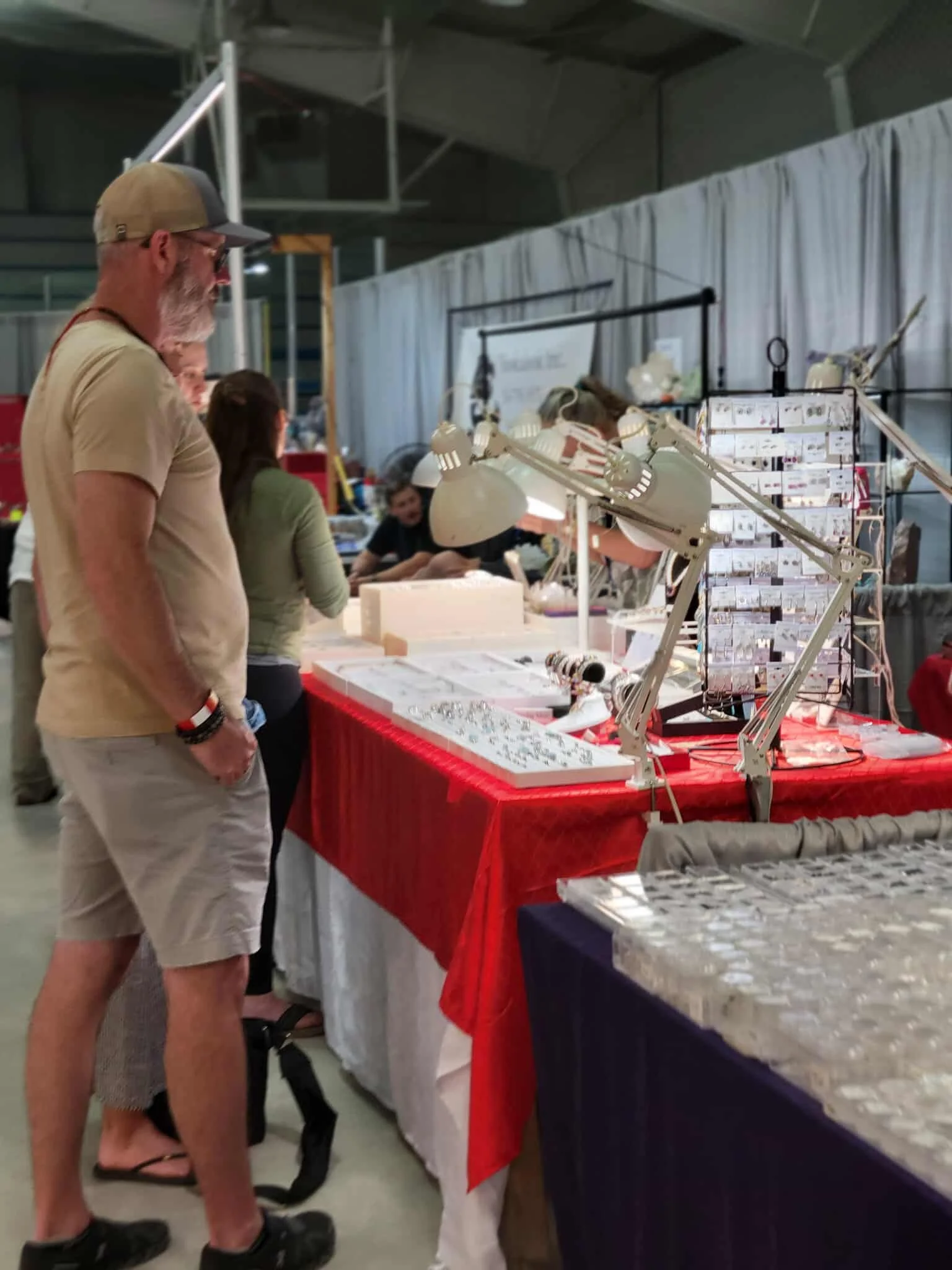 A man with a baseball cap and beard looking at jewelry displayed on a table at a market or craft fair. The jewelry is arranged on display racks with lights, and there are other people behind the table.