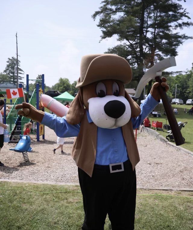 Person in a Pluto costume dressed as a Canadian Mountie holding a pickaxe at a park or playground with children and playground equipment in the background.