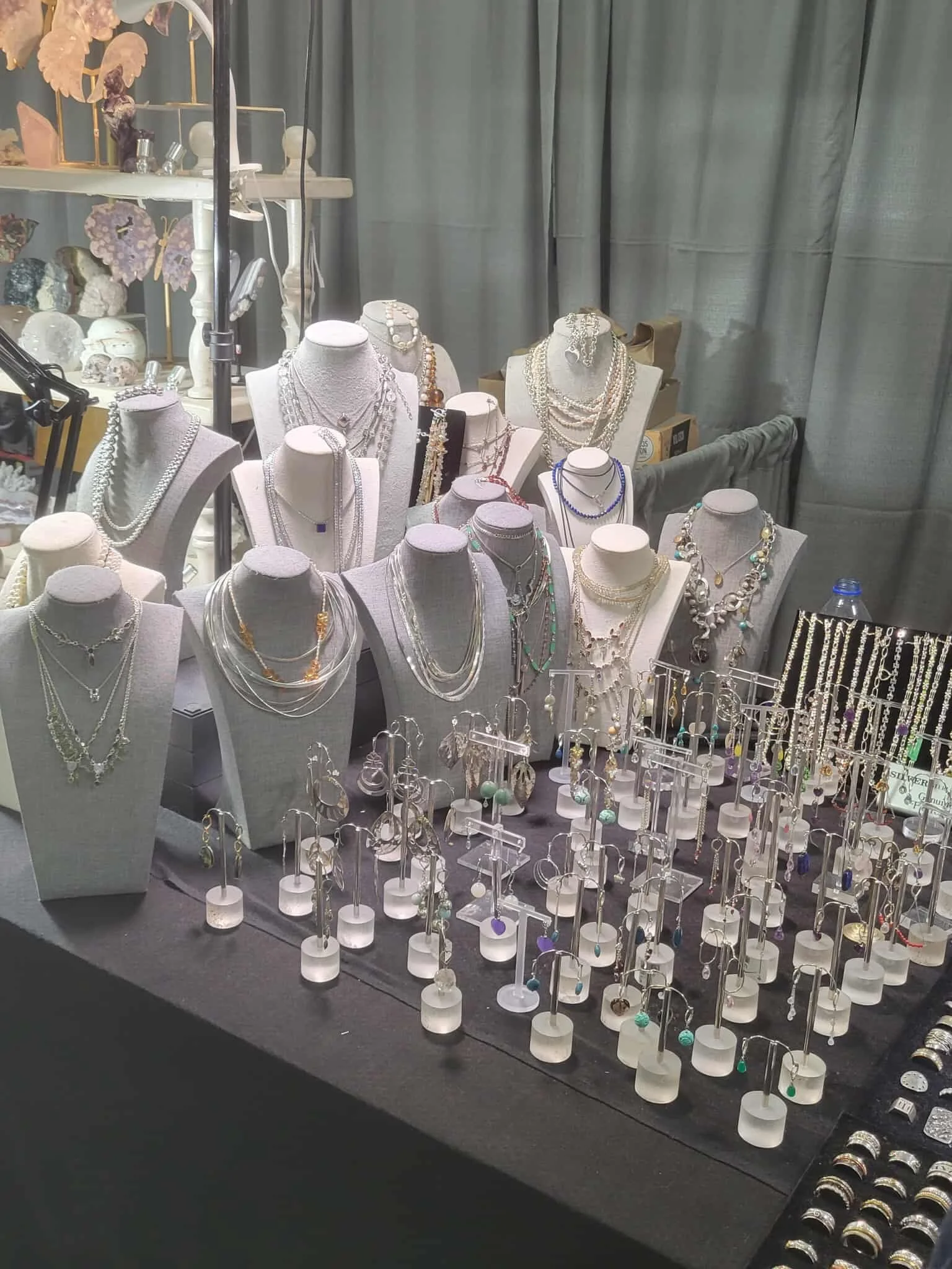 Display of various jewelry items, including necklaces, earrings, and rings, arranged on white busts and small stands at a jewelry booth, with a backdrop of grey curtains.