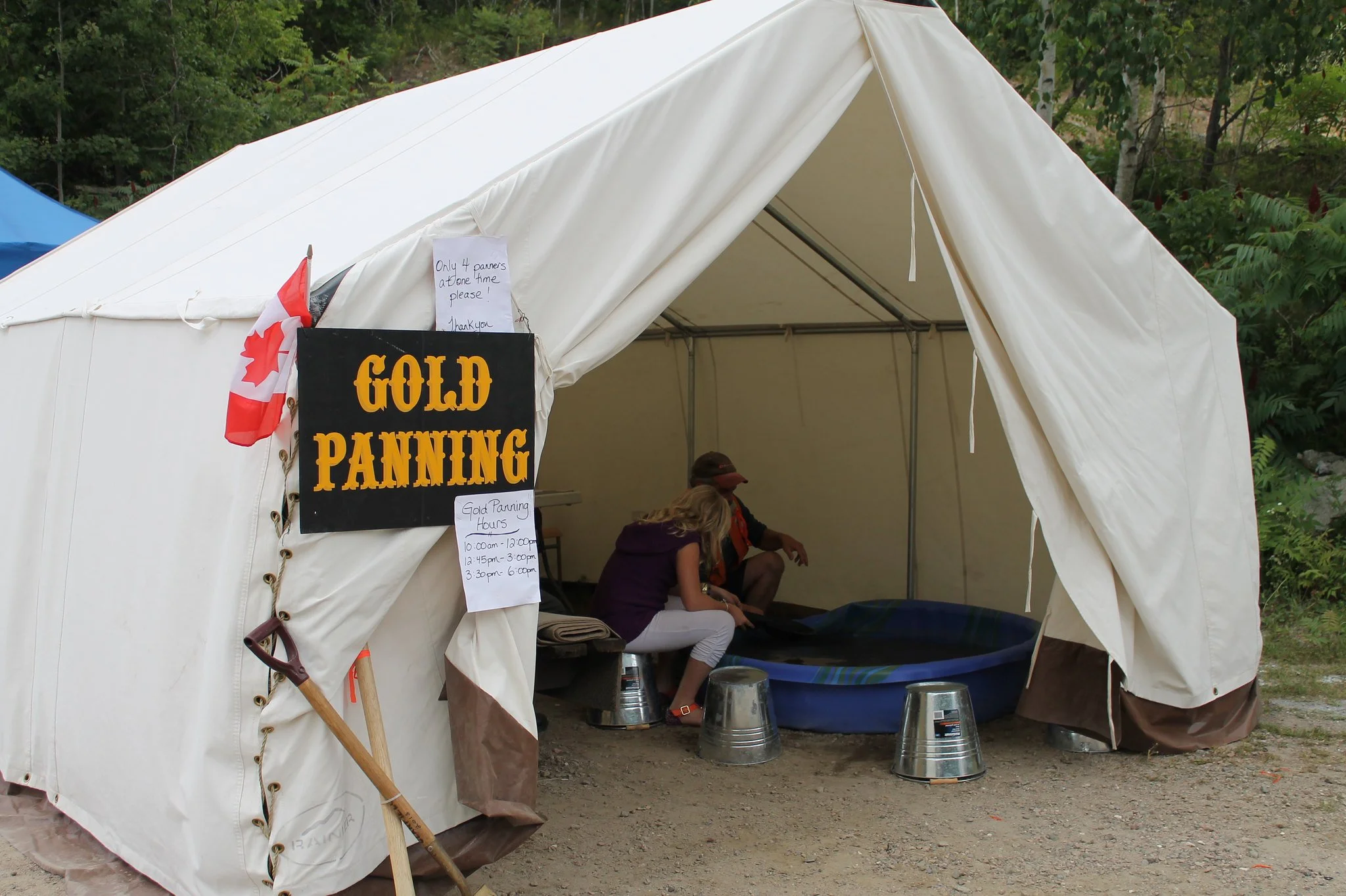 A large white tent with a sign that says 'Gold Panning' at an outdoor event, with trees in the background and people sitting inside.