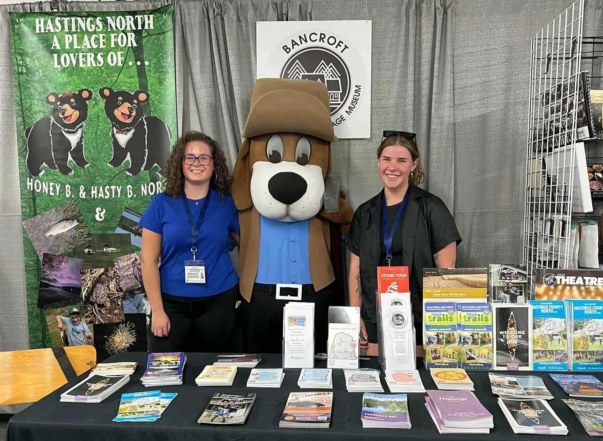Booth at a fair or exhibition featuring banners with forest and wildlife images, brochures, and a mascot costume of a dog wearing a hat, with two young women standing next to and in front of the mascot.