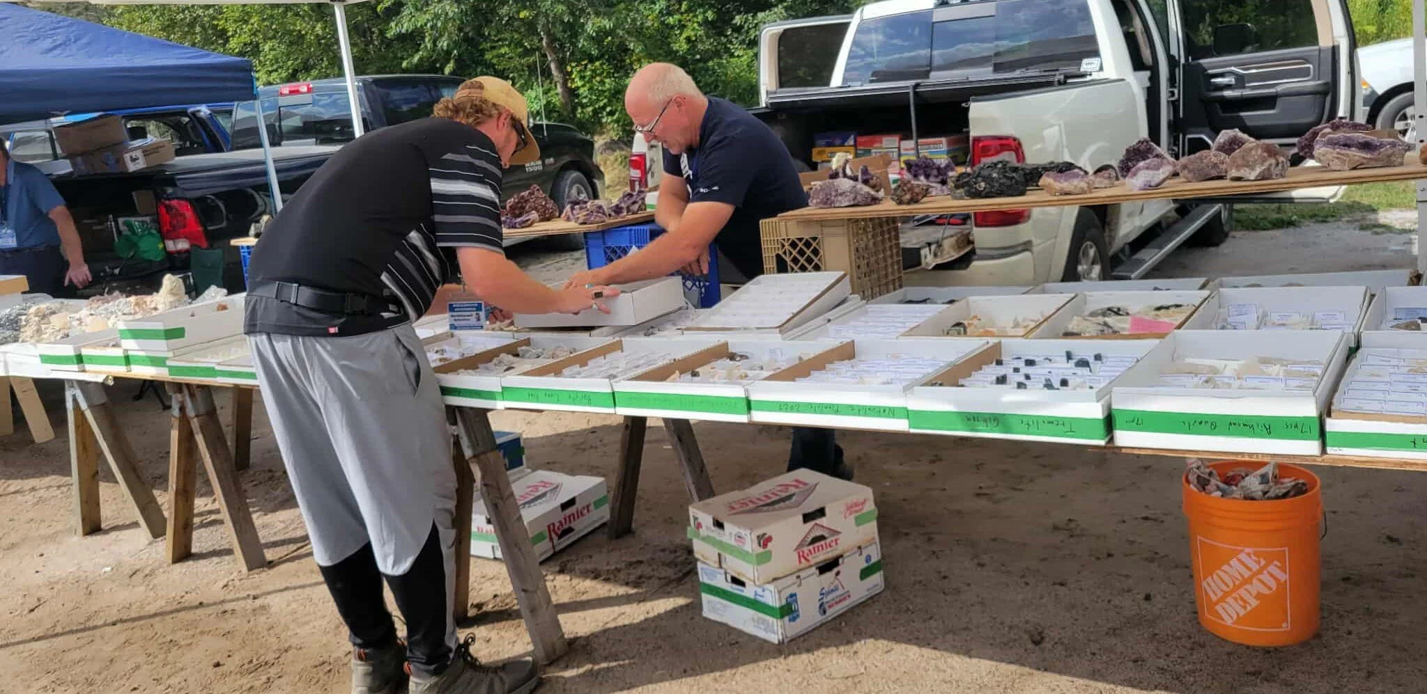 Two men at an outdoor gemstone sale, examining rocks and minerals displayed on tables under a canopy, with cars parked in the background.