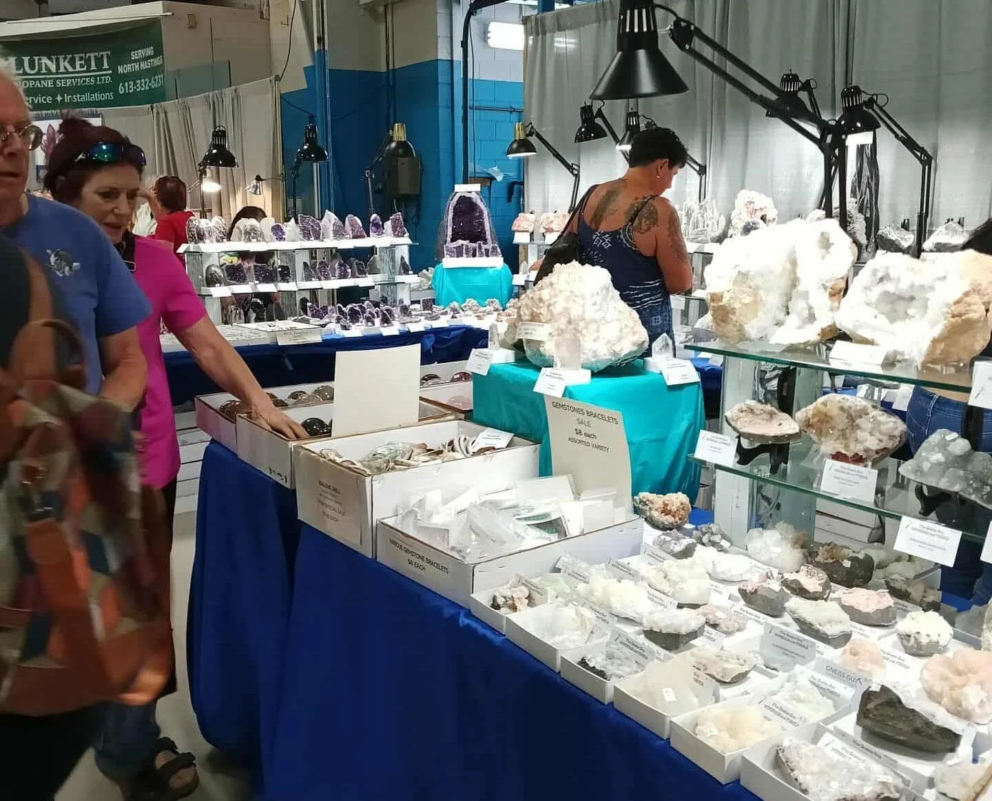 People shopping at a mineral and crystal market, with various rocks, minerals, and crystals displayed on tables and shelves, indoors with industrial lighting.