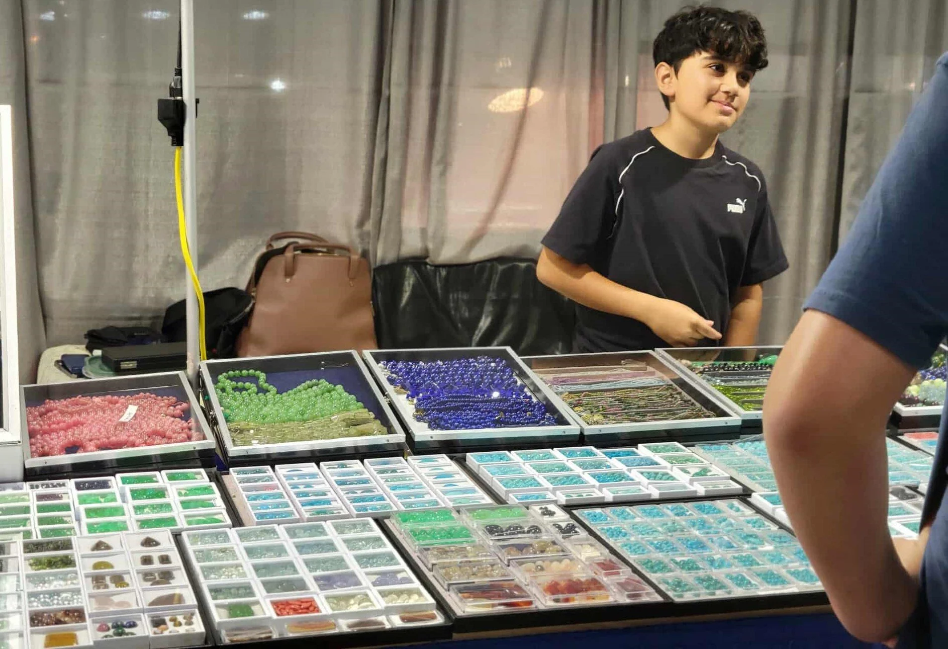 A boy in a black Puma t-shirt standing at a jewelry booth with trays of colorful beads and jewelry supplies at a craft fair or market.