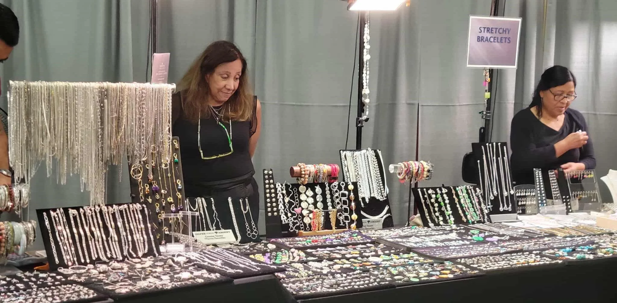 Jewelry display booth with various necklaces, bracelets, and earrings, with two women tending the booth at a craft fair.