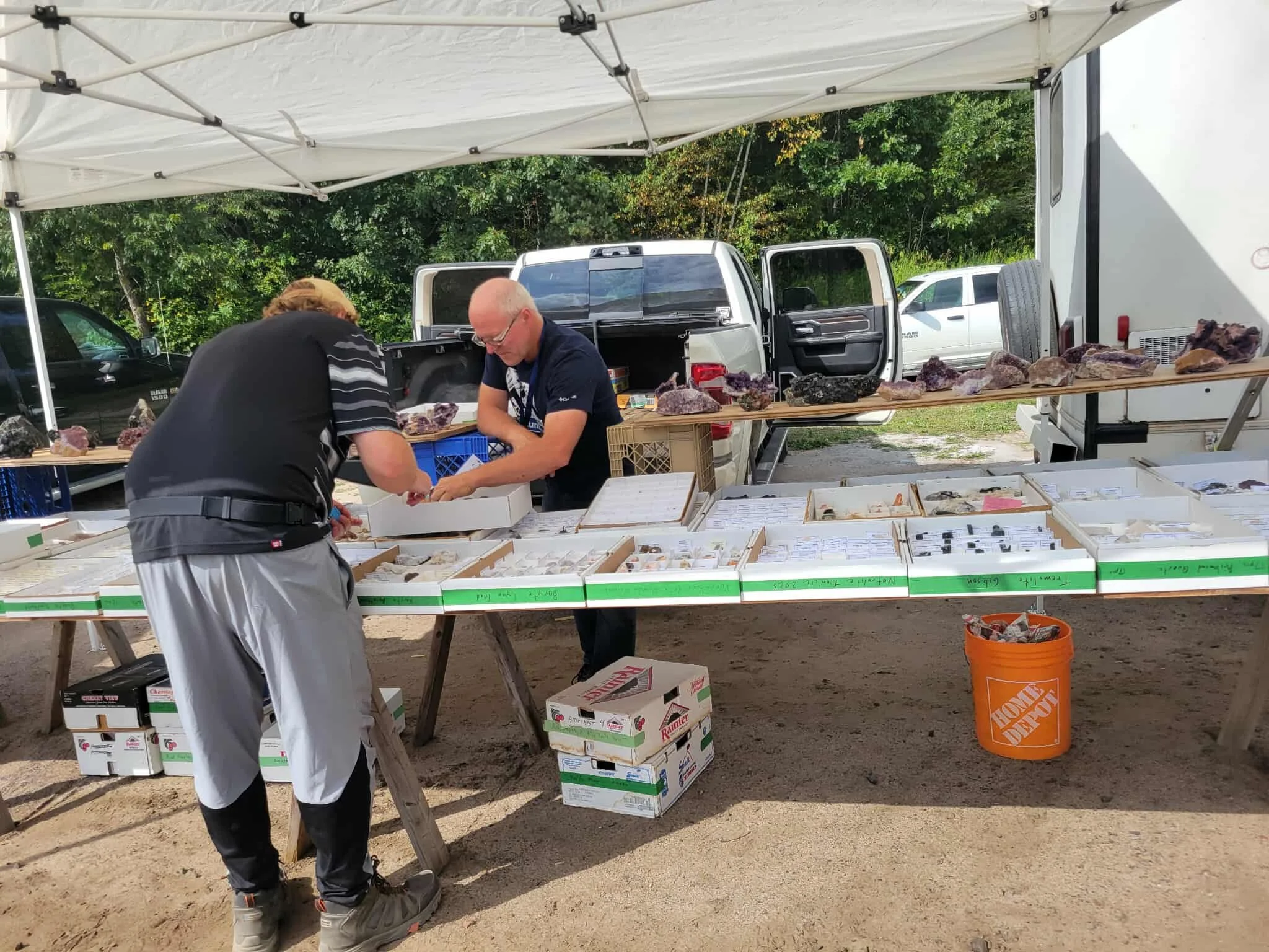 Two men at an outdoor booth examining rocks or minerals displayed on tables, with boxes underneath and a tent overhead.