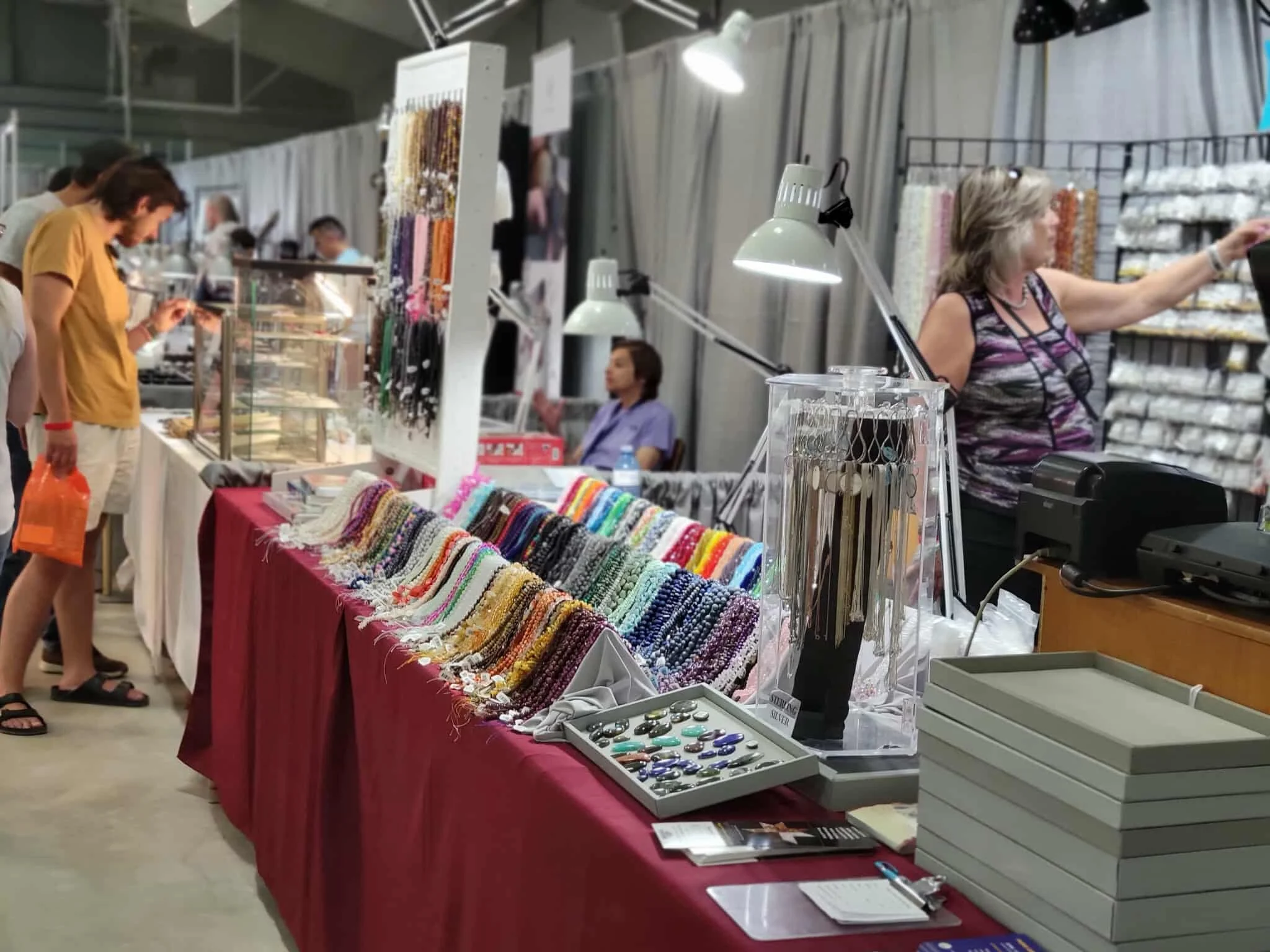 People shopping at a craft fair or market, with tables of jewelry, buttons, and fabric crafts on display.