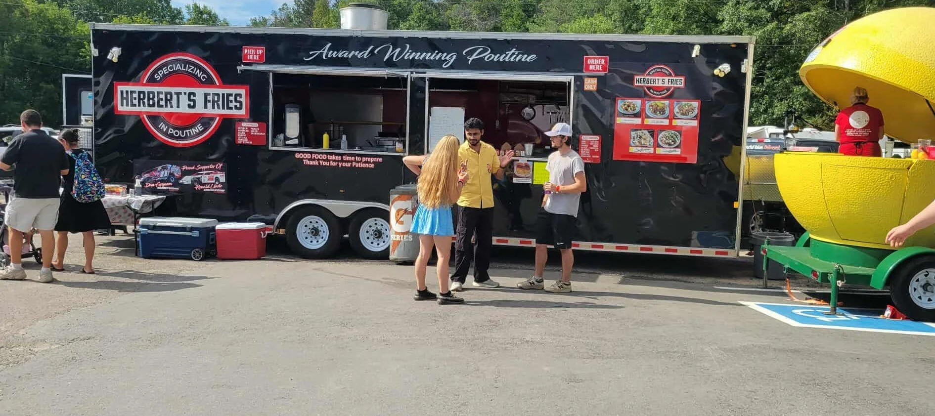 A food truck named 'Herbert's Fries' serving poutine, with a group of people ordering and chatting in front of it. There's a yellow container on a green cart on the right side and some coolers on the ground. The scene appears to be at an outdoor even