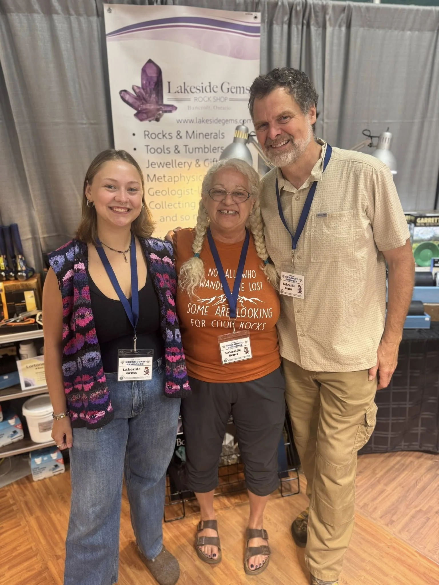 Three people standing together in front of a Lakeside Gems banner at a gemstone event, smiling at the camera. The person in the middle is wearing glasses, a brown t-shirt, and sandals, with braided hair. The person on the left is wearing a colorful c
