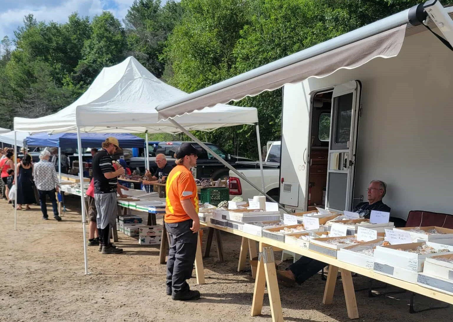 An outdoor marketplace with vendors selling baked goods from a trailer and tables, under white canopies, with people browsing and buying items in a park or rural area with trees in the background.