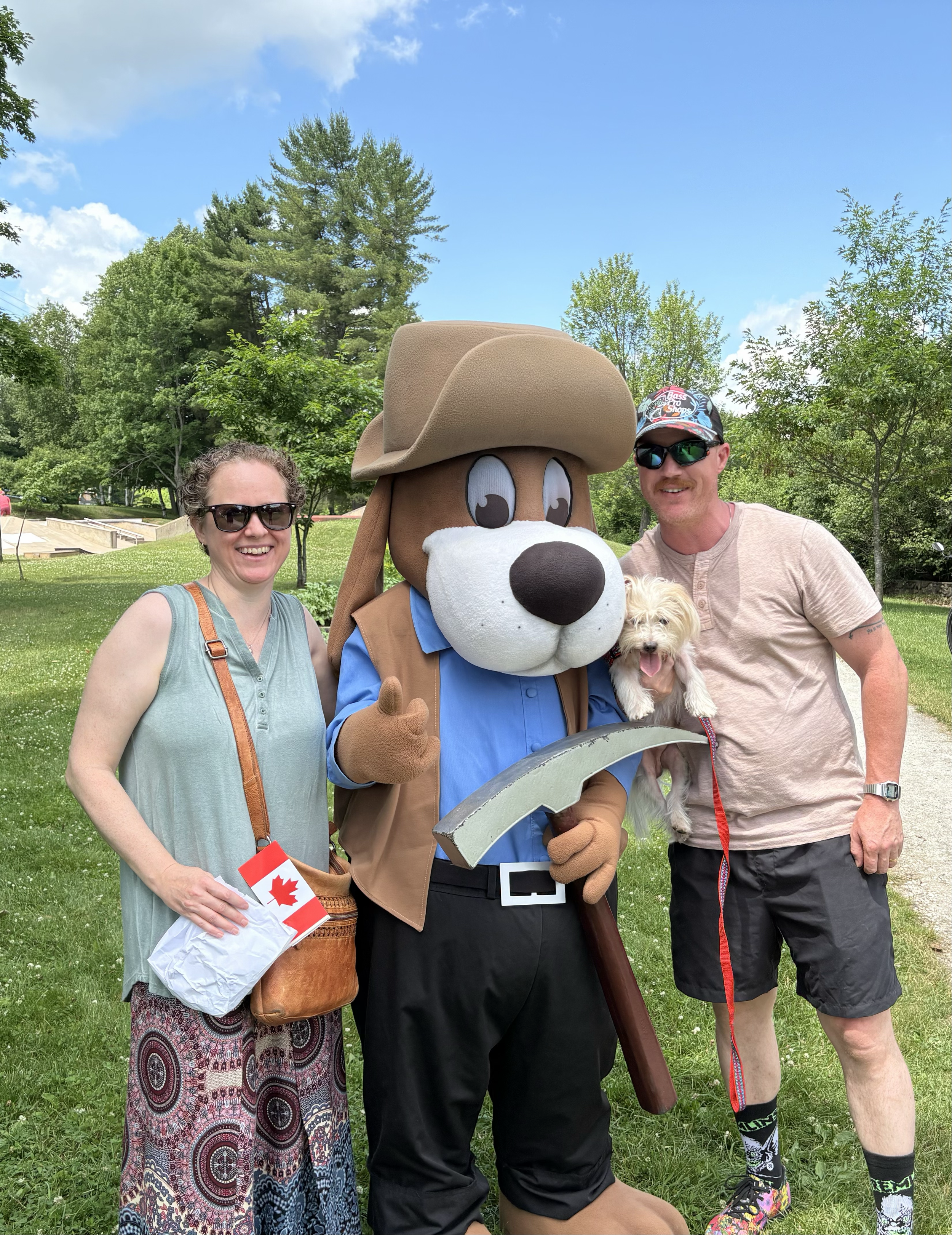Two women and a man standing outdoors on grass with trees in the background. The man is in a mascot costume of a dog wearing a large cowboy hat, with a person in casual clothes holding a small dog. One woman is holding a Canadian flag.