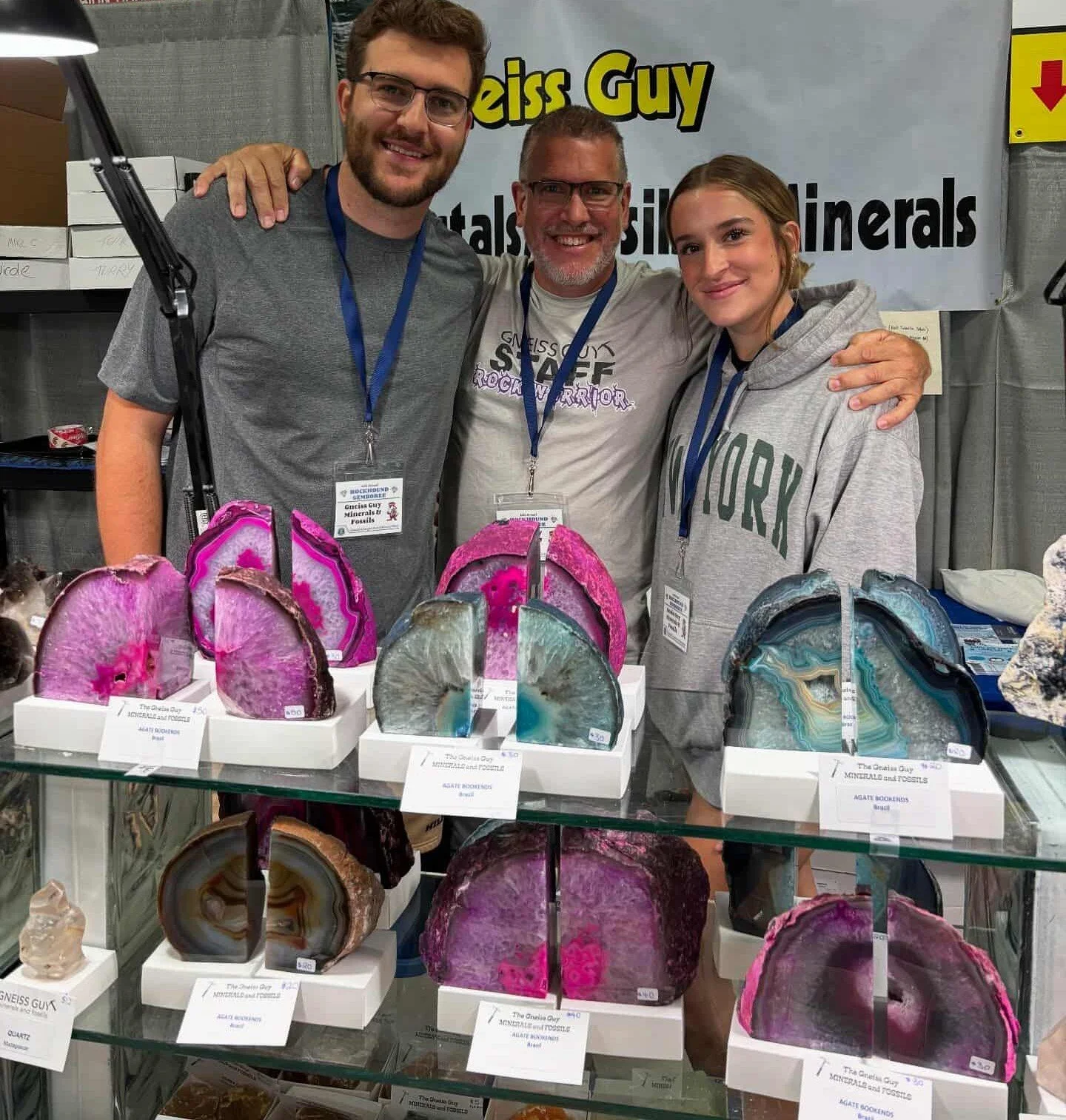 Three people smiling and posing at a mineral and gemstone exhibit, with various colorful agate and quartz slices on display in front of them and a banner in the background.