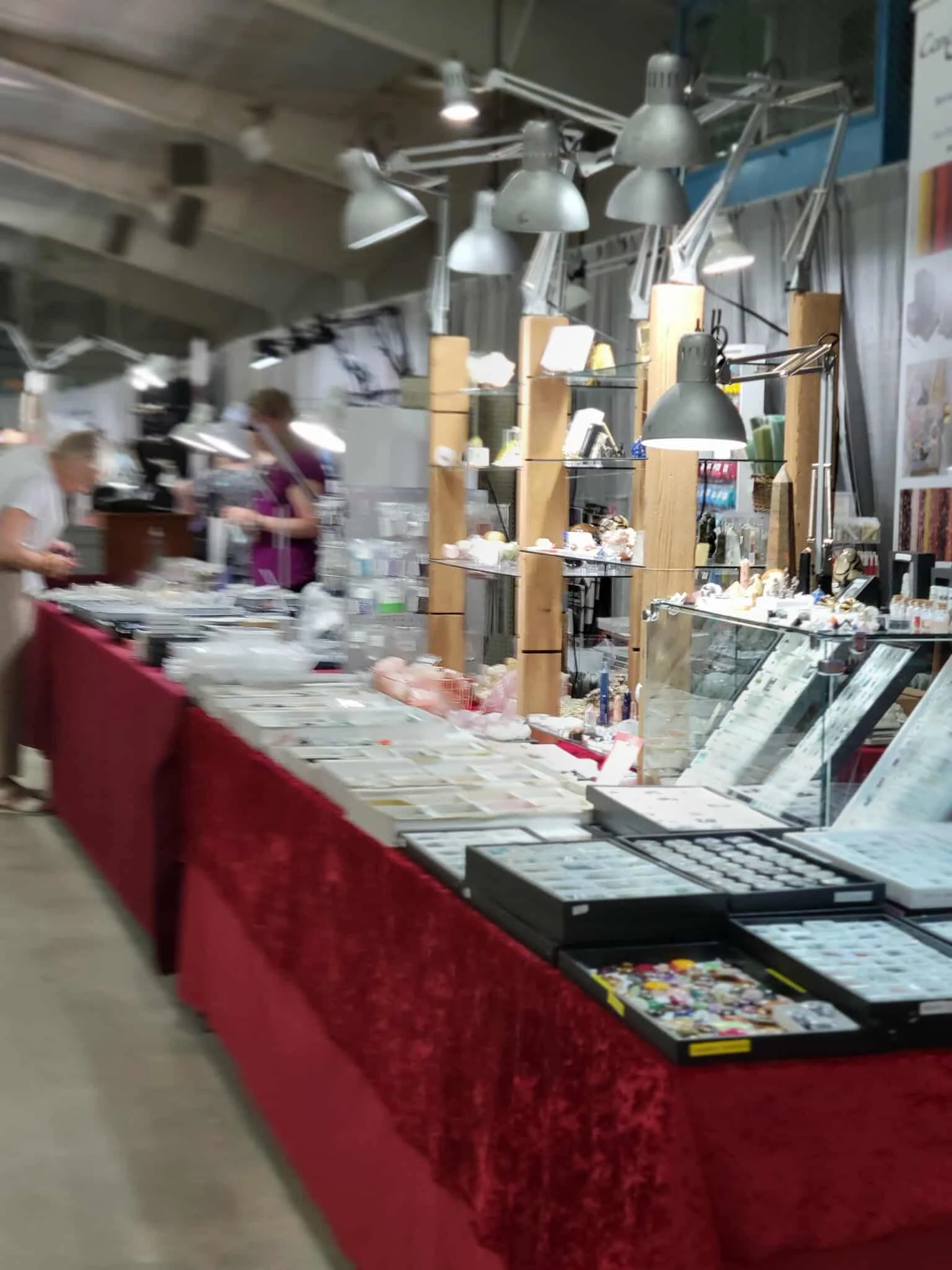 A display table at a market or fair with various jewelry and small decorative items, covered with a red cloth, with glass shelves for additional items, and silver adjustable industrial-style lamps overhead, in an indoor setting with a fabric curtain 